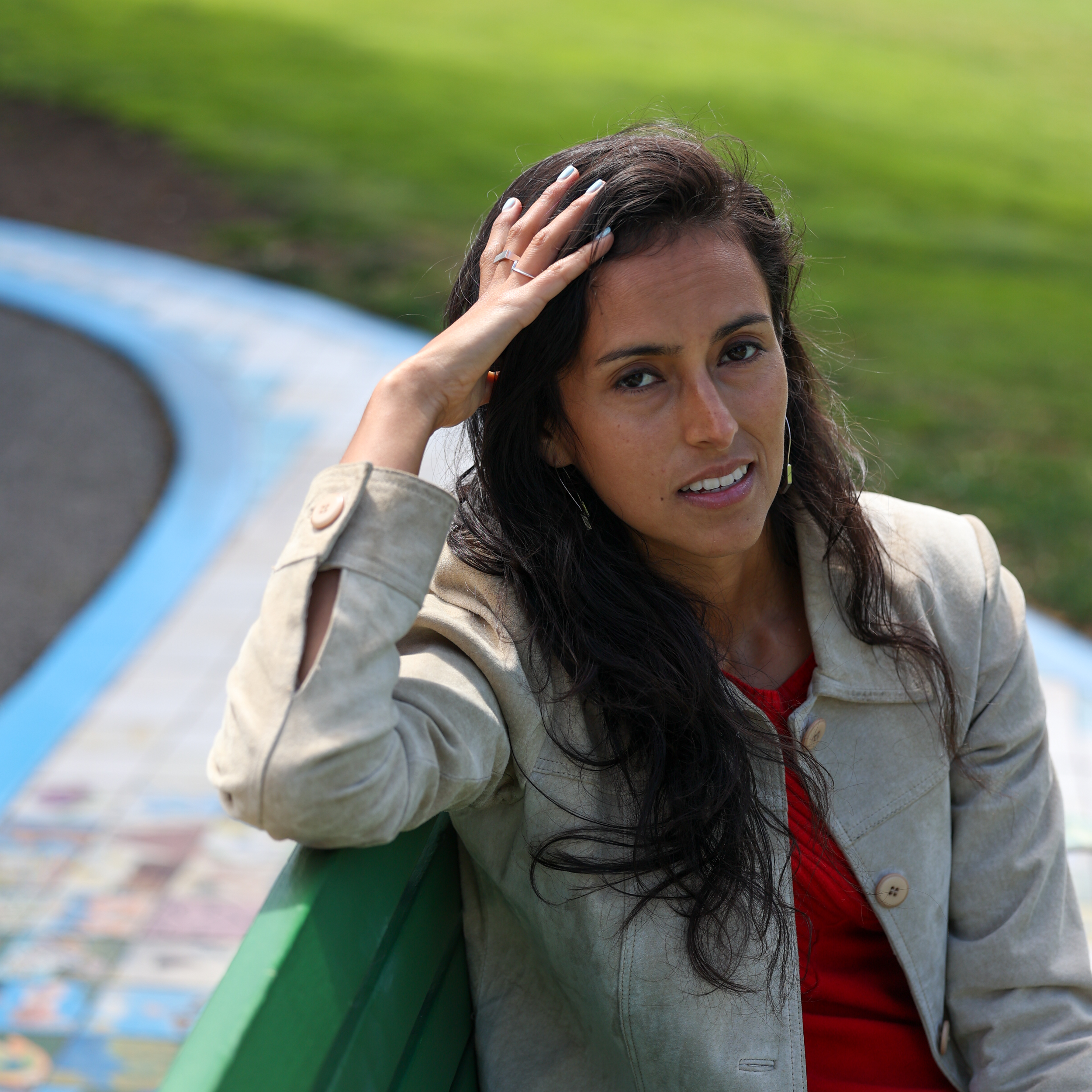 A woman with long, dark hair sits on a green bench, wearing a beige jacket and red shirt. She rests her hand on her head with a grassy background behind her.