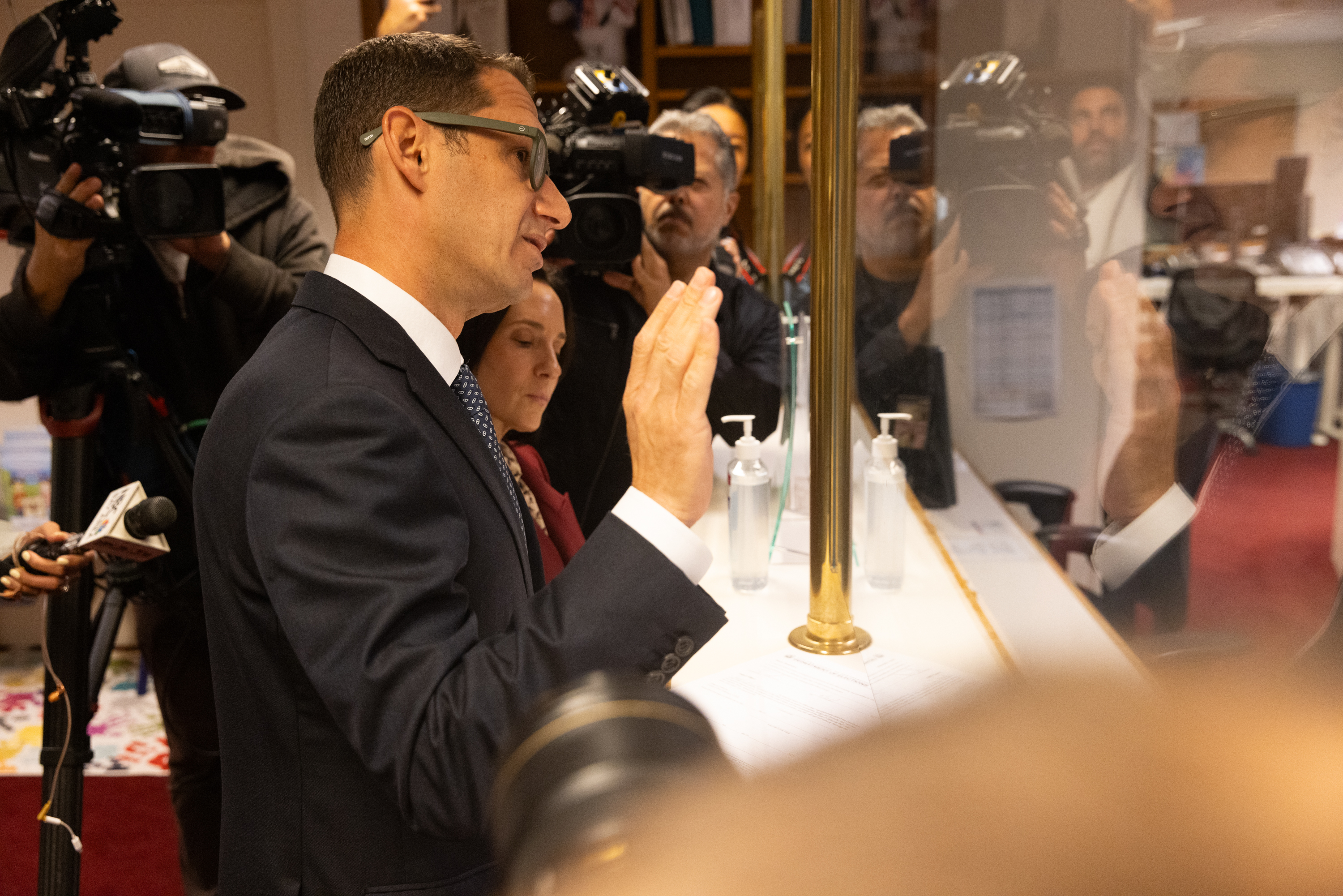 A man in a suit speaks behind a counter with cameras and reporters capturing the scene. Two bottles of sanitizer are visible next to him.