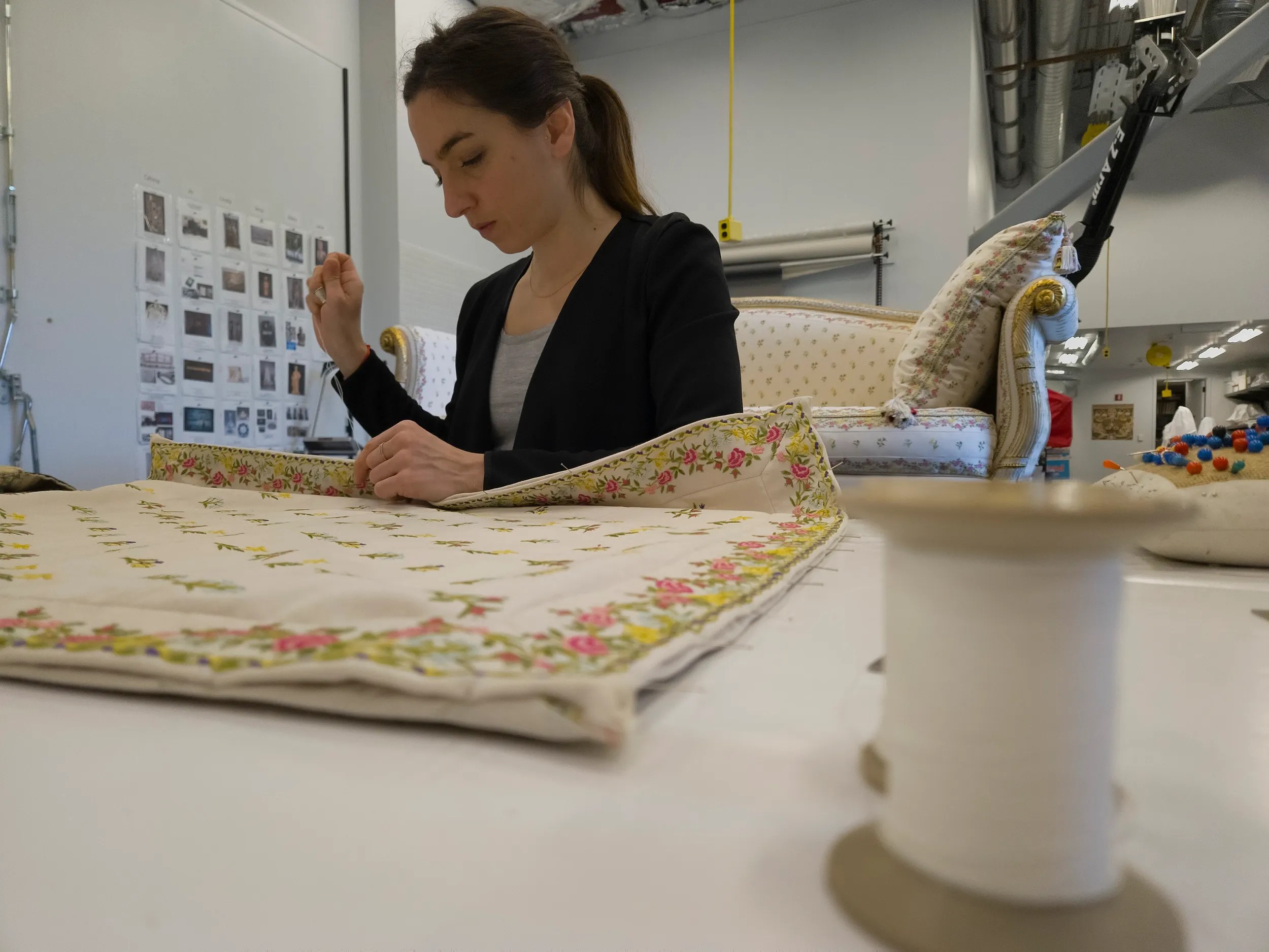 A woman in a workshop is focused on sewing a floral-patterned fabric. A spool of thread and pincushion are on the table, with a vintage sofa in the background.