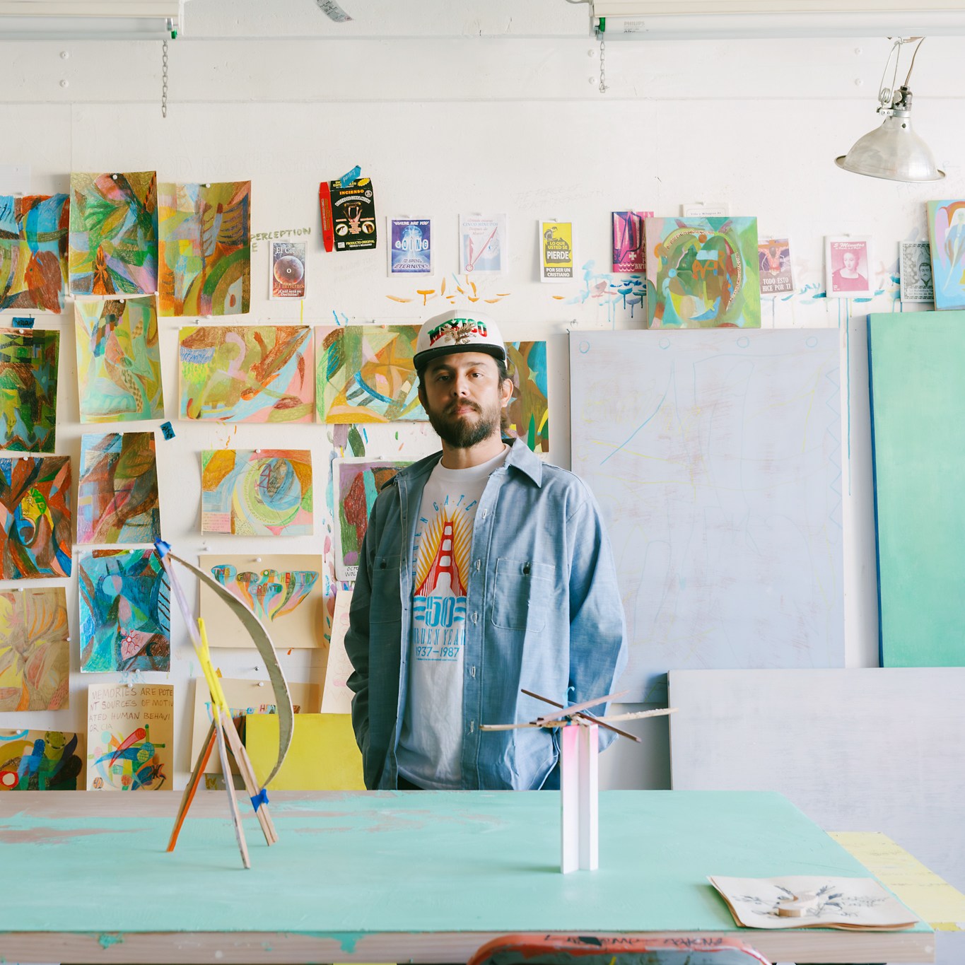 A bearded man in casual clothing stands in an art studio with colorful abstract paintings covering the walls. A sculpture sits on a table in front of him.