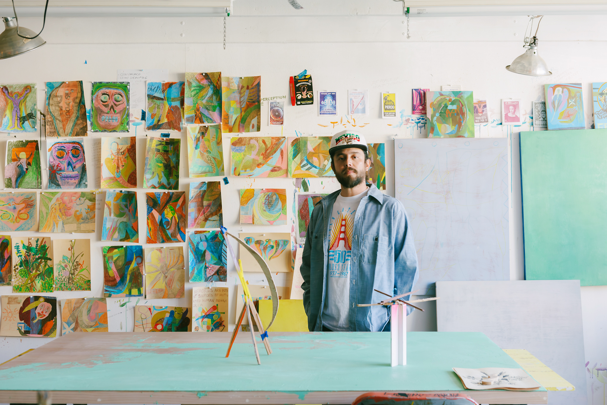 A bearded man in casual clothing stands in an art studio with colorful abstract paintings covering the walls. A sculpture sits on a table in front of him.