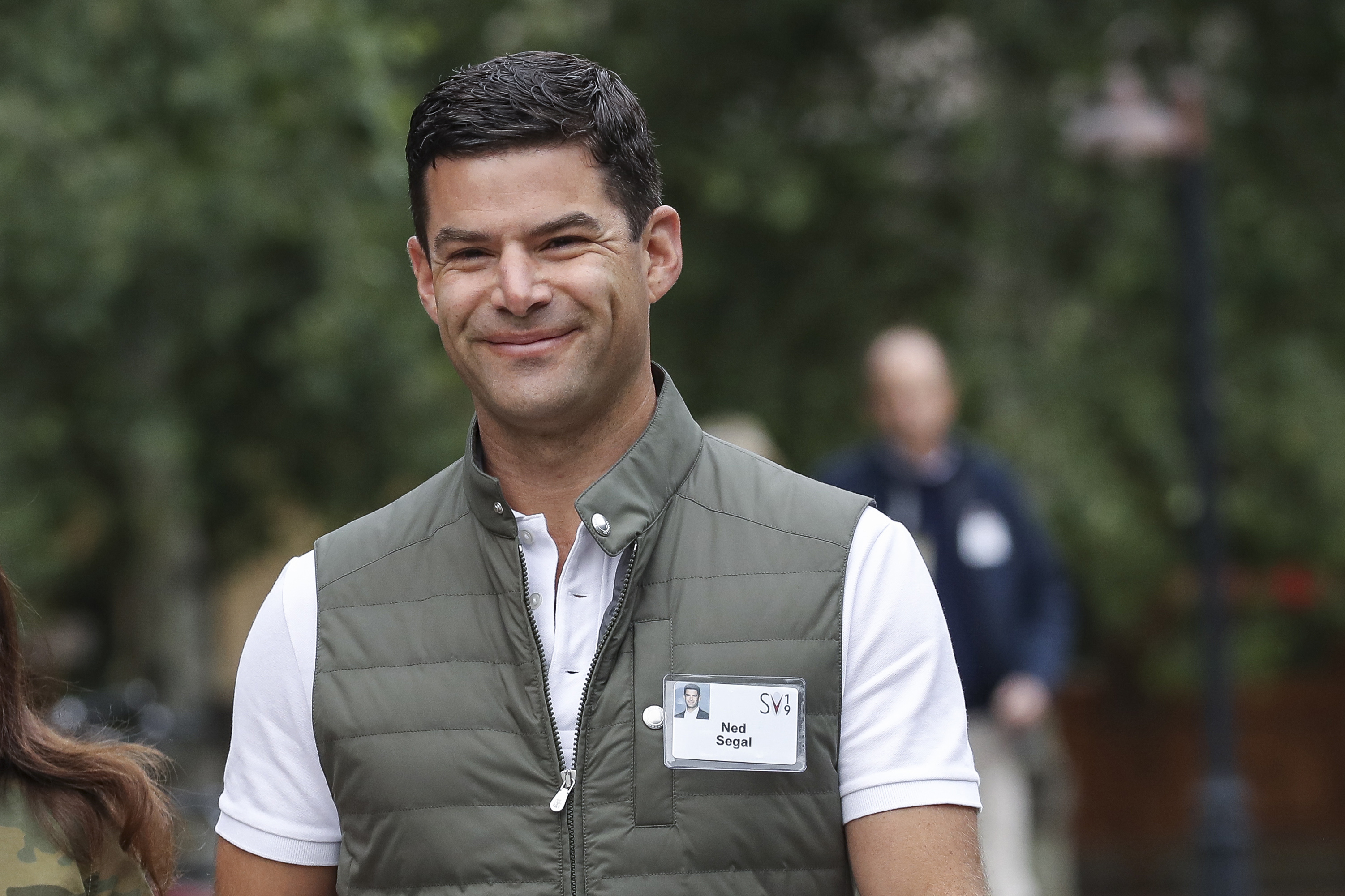 A person is smiling outdoors, wearing a white shirt and a green vest with a name tag. The background is blurred and has greenery.