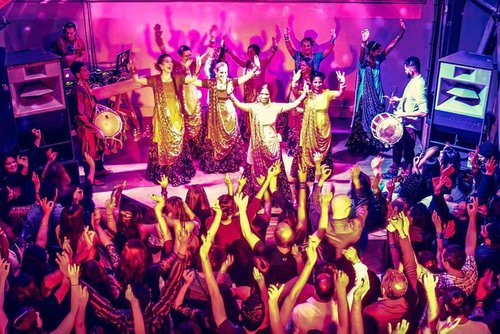 A lively dance performance shows a group on stage in colorful, traditional attire, raising their arms. They are surrounded by an enthusiastic crowd below, also raising hands.