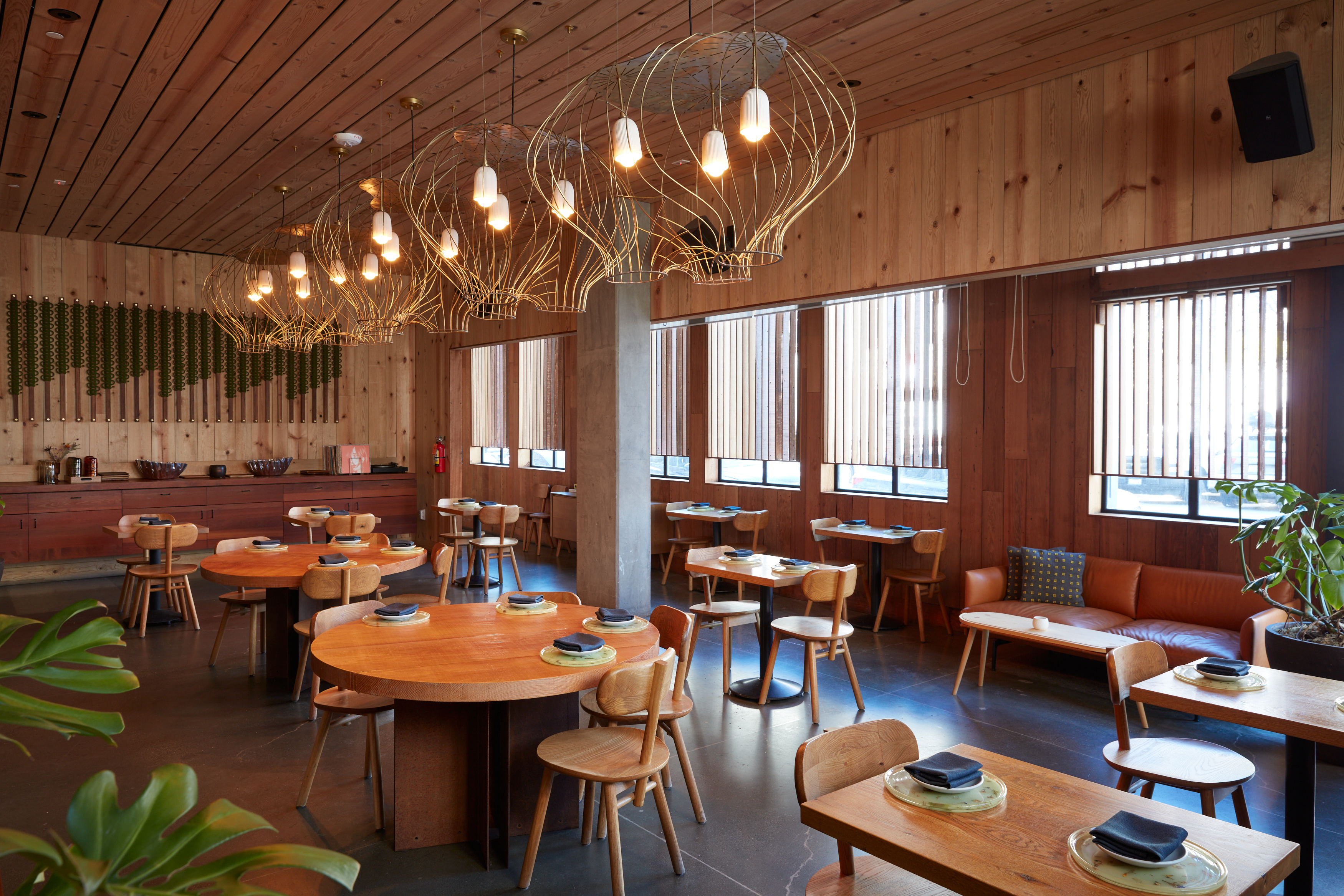 The image shows a stylish restaurant with wooden furniture, round tables, pendant lights, and large windows. A couch is along one wall, adding a cozy touch.