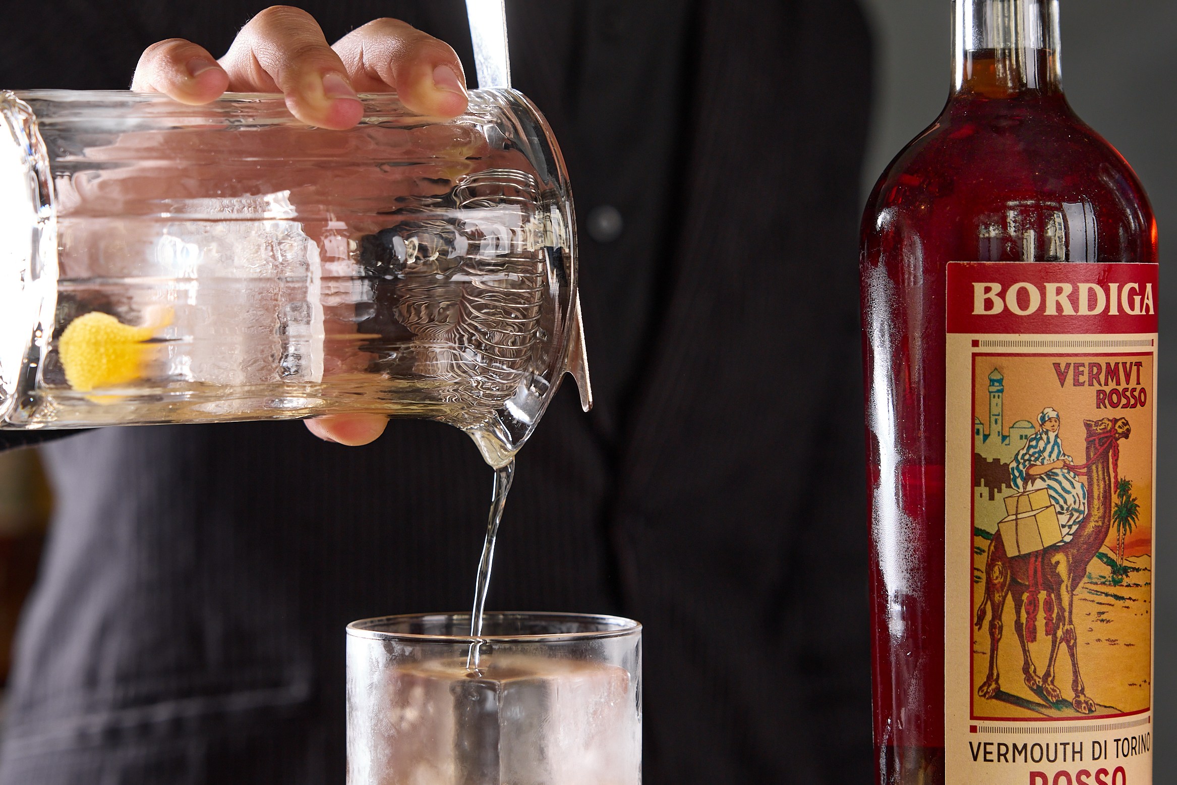 A person in a suit pours clear liquid from a glass pitcher into a glass with ice. A bottle of Bordiga Vermouth Rosso stands on the wooden table beside them.