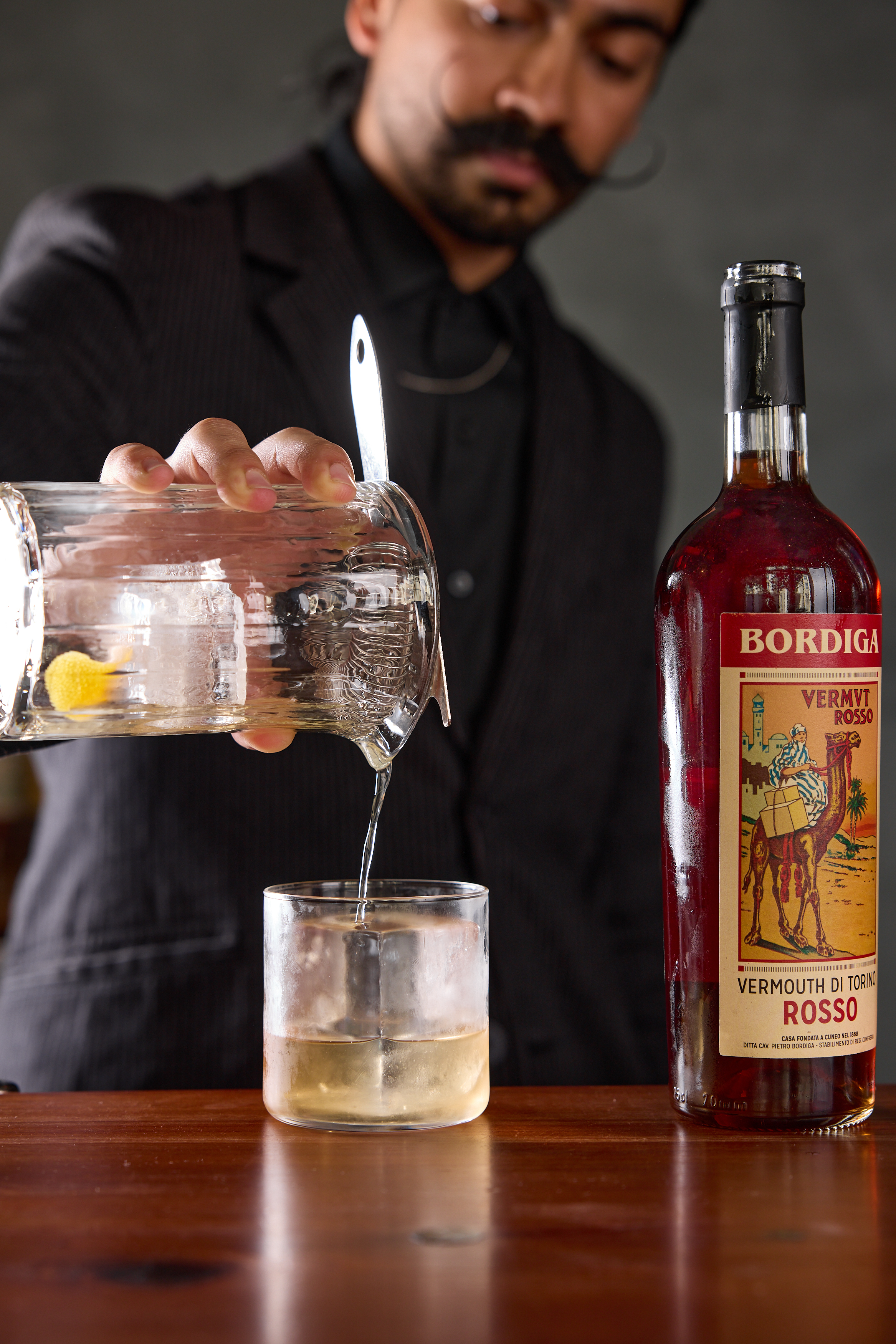 A person in a suit pours clear liquid from a glass pitcher into a glass with ice. A bottle of Bordiga Vermouth Rosso stands on the wooden table beside them.