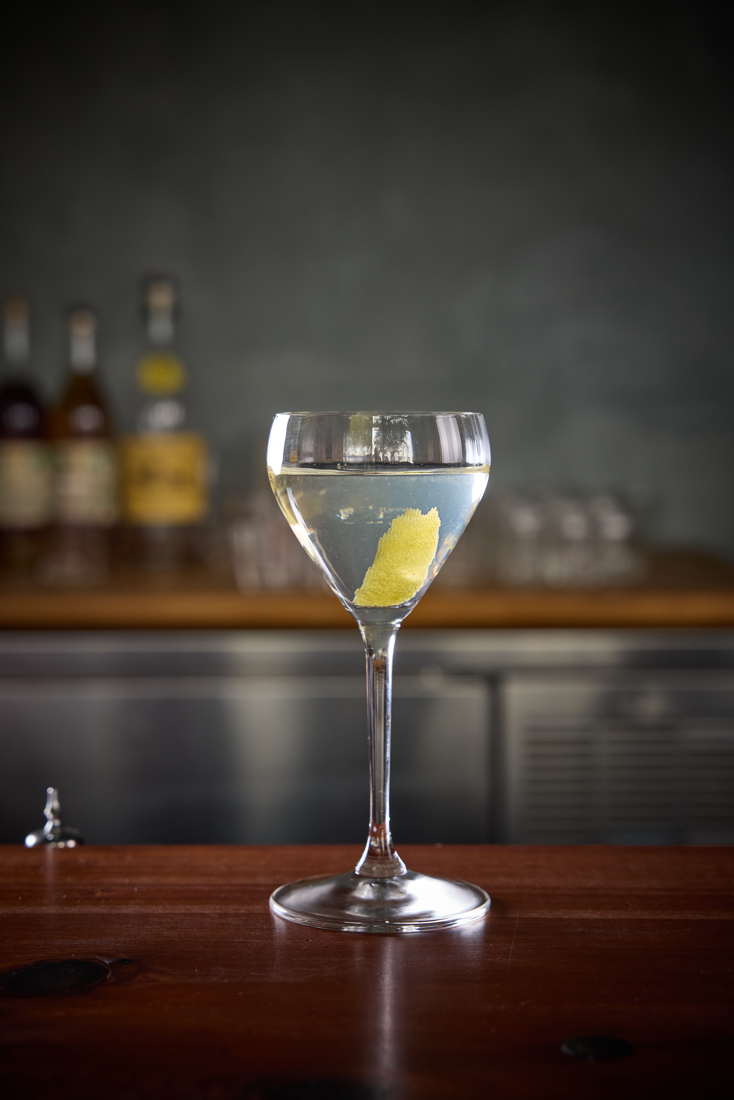 A cocktail glass sits on a wooden bar with a clear drink and a lemon twist inside. Blurry bottles and glasses are visible in the soft-focus background.