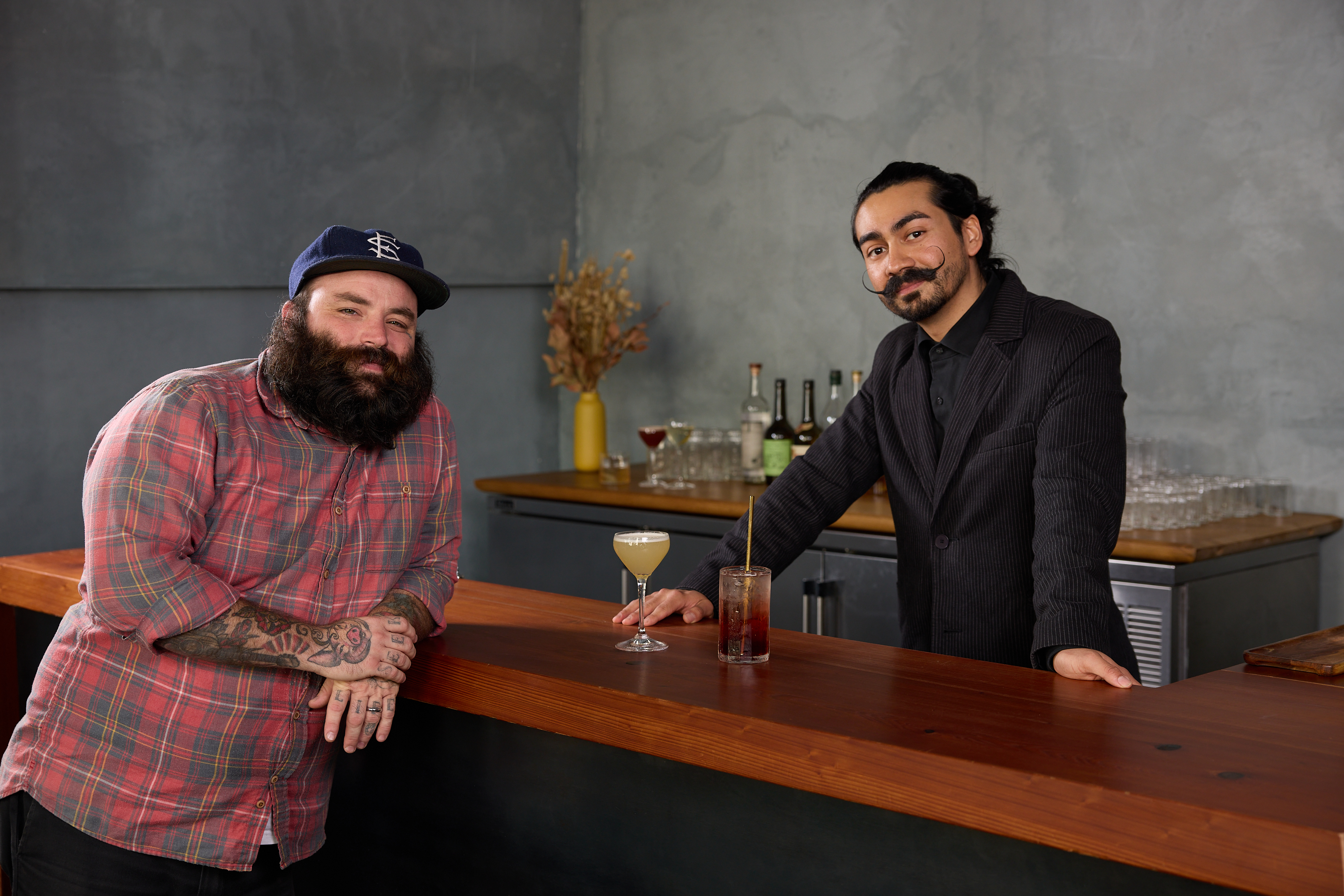 Two men stand behind a wooden bar. One is in a plaid shirt and hat, the other in a black suit. Cocktails and bottles are on the bar, with a gray wall backdrop.