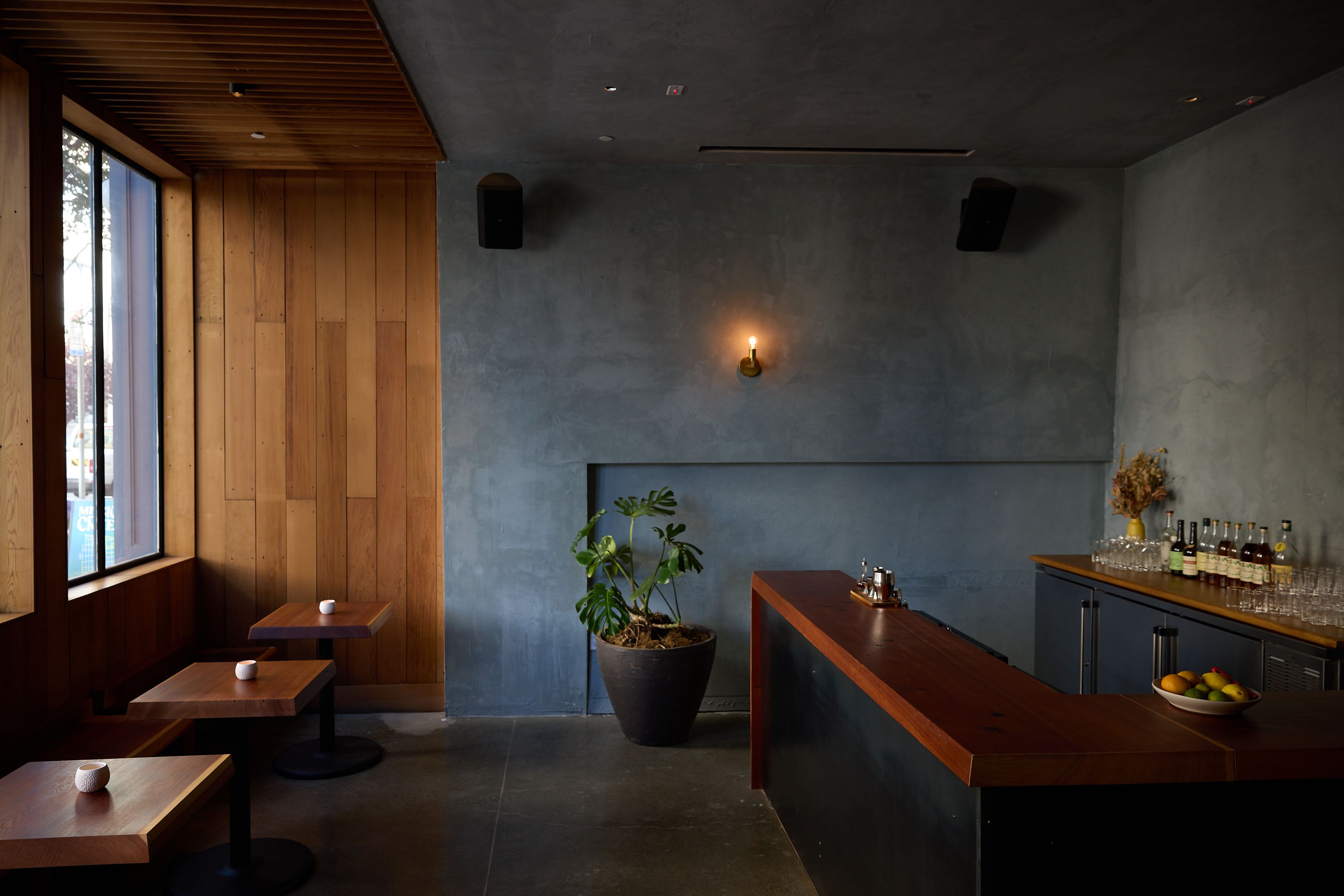 A cozy bar with wooden tables and chairs, a wooden counter, a potted plant, dim lighting, and bottles on display. The walls are dark with a single sconce.