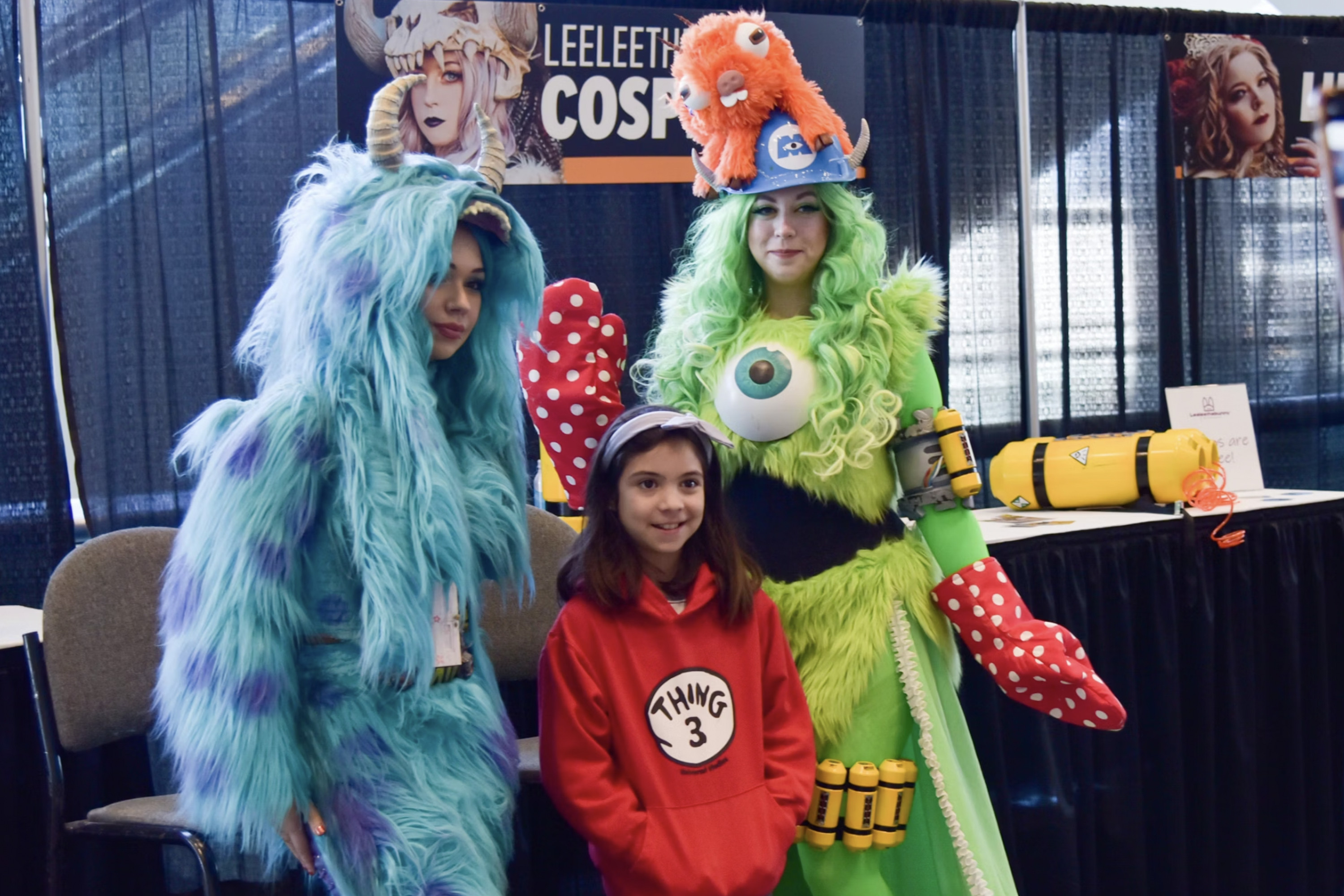 A girl in a red hoodie poses with two adults in colorful, furry monster costumes with horns and big eyes at a cosplay event.