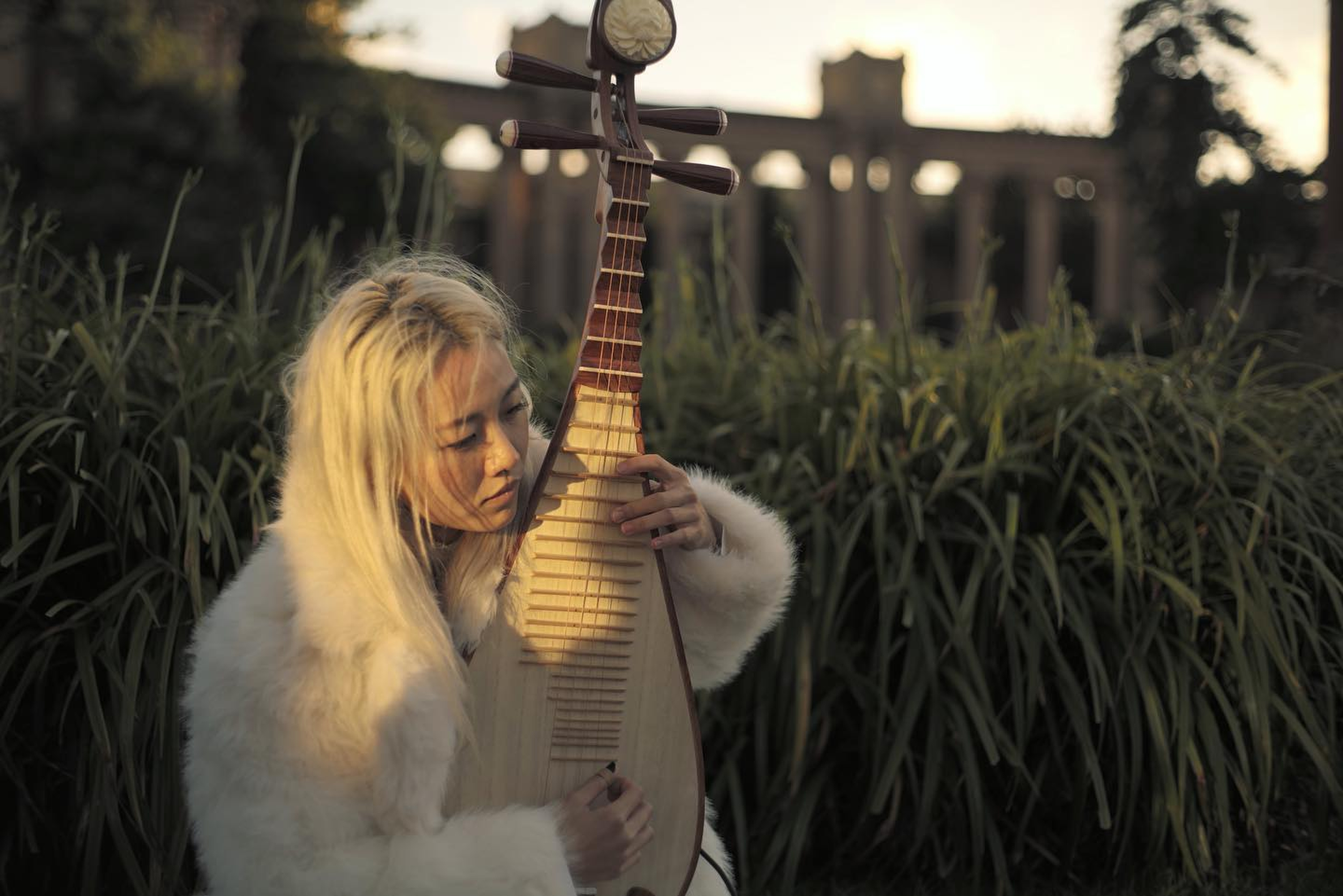 A blonde-haired person in a white fluffy coat plays a stringed instrument outdoors, with tall grass and a large concrete structure in the background.