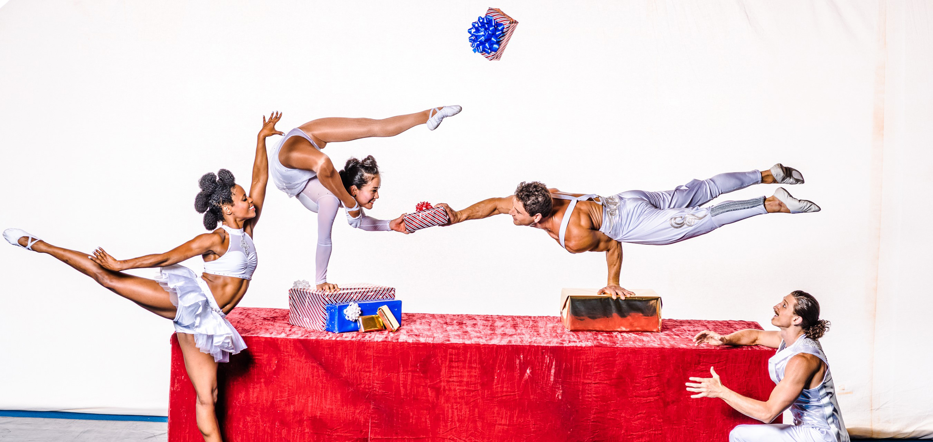 Four acrobats in white perform dynamic poses on and around a red table, interacting with gift-wrapped presents, showcasing balance and agility.