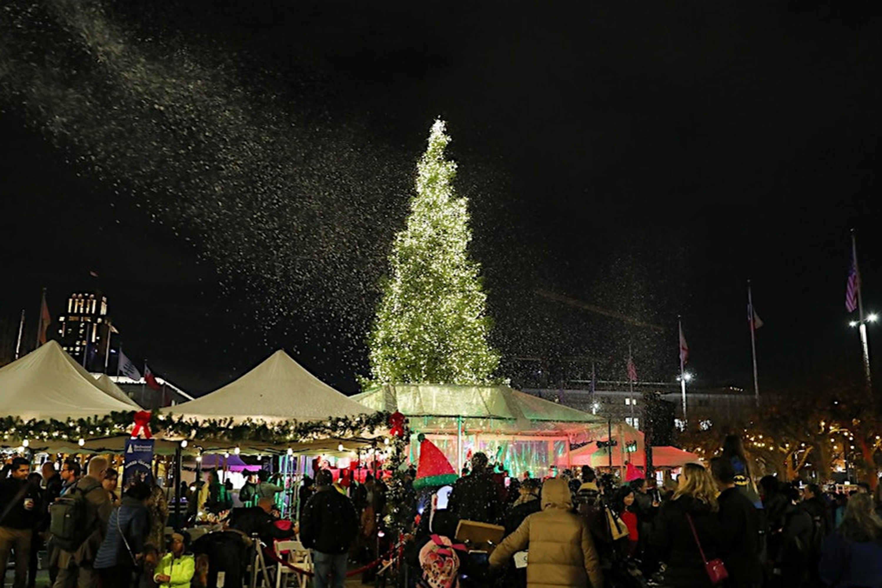 The image shows a festive crowd under a large, illuminated Christmas tree with fake snow falling. People are gathered around decorated tents at night.