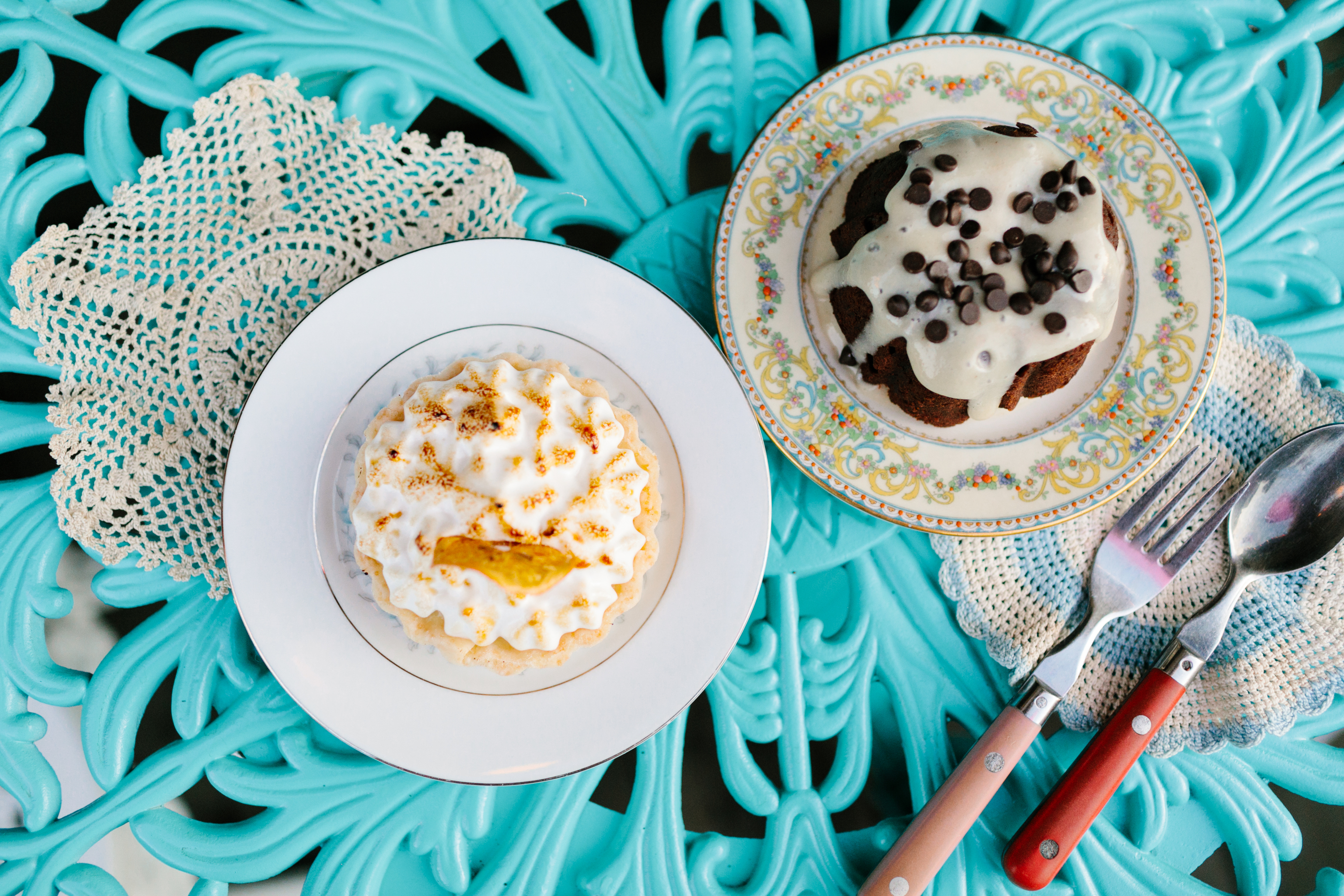 Two desserts sit on a turquoise table, one with whipped cream and a lemon slice, the other with chocolate chips, accompanied by colorful cutlery.