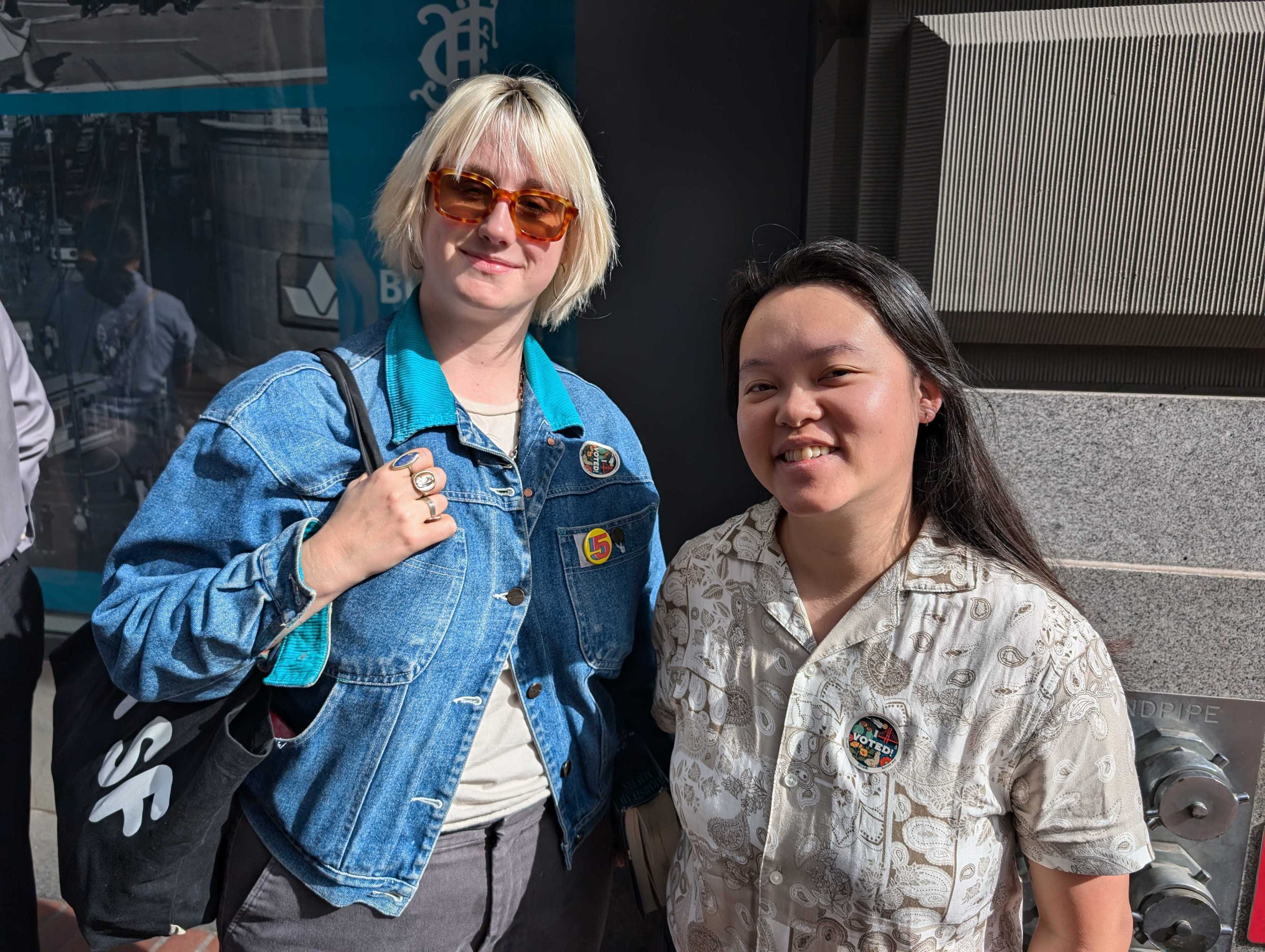 Two people stand outside, smiling. One wears a denim jacket and sunglasses, holding a bag. The other wears a patterned shirt with an &quot;I Voted&quot; sticker.