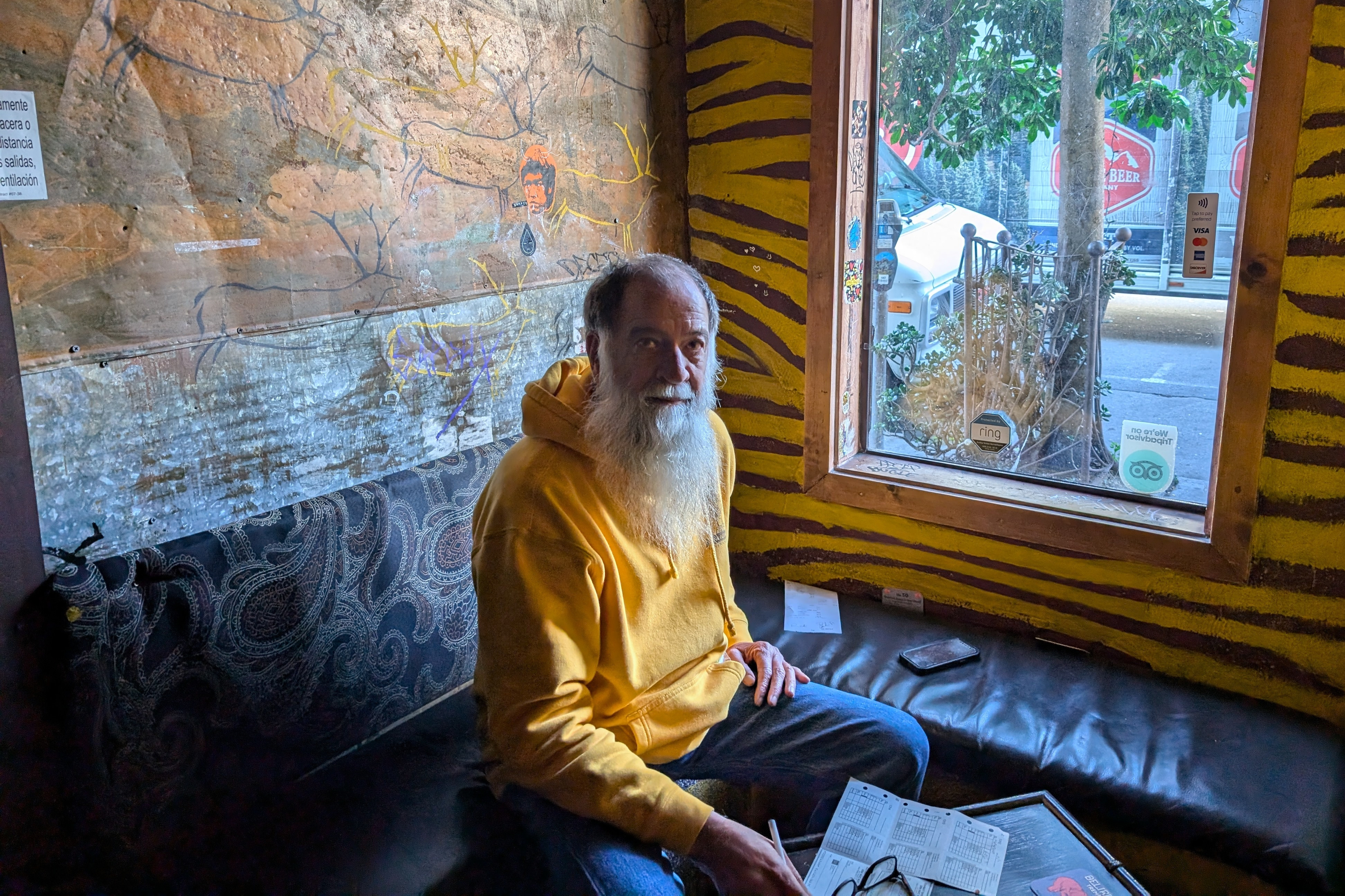 A man with a long beard in a yellow hoodie sits on a patterned couch next to a window. He holds a newspaper and looks at the camera, surrounded by eclectic decor.