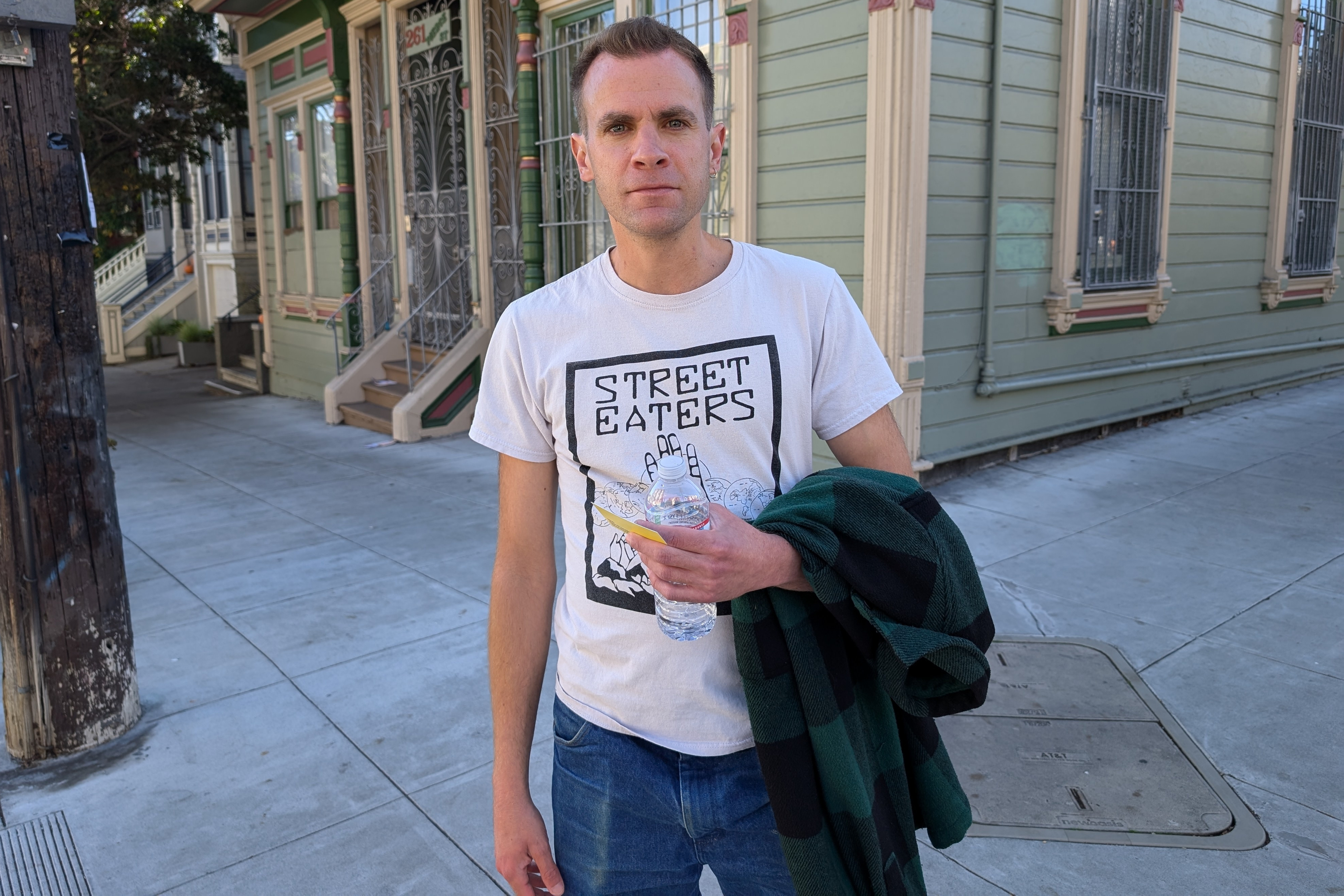 A person wearing a &quot;Street Eaters&quot; t-shirt holds a bottled drink and a green plaid jacket. They stand on a sidewalk beside buildings with ornate trim.