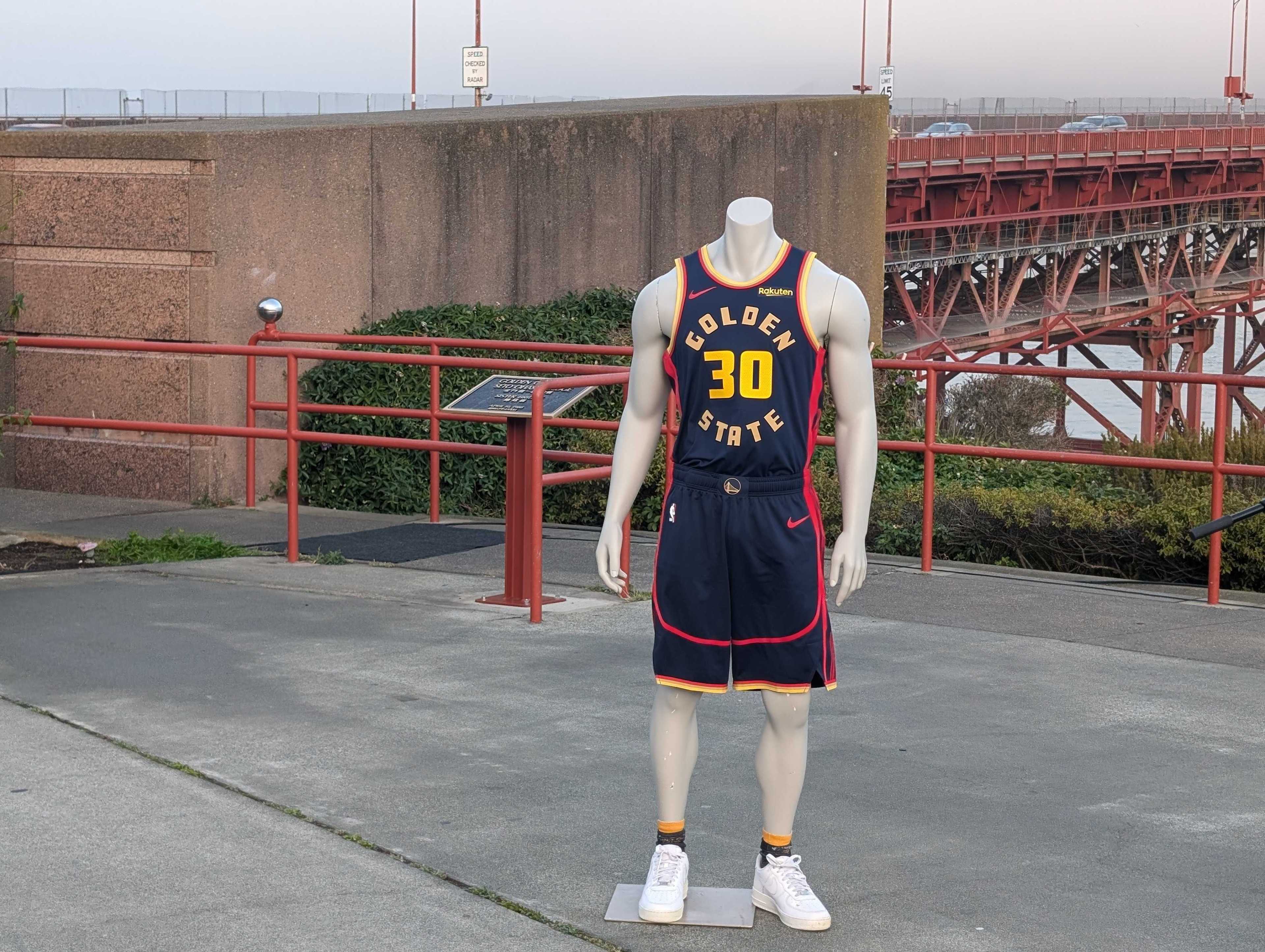 A mannequin wearing a Golden State Warriors basketball uniform faces forward. It's outdoors with a view of a bridge and some greenery in the background.