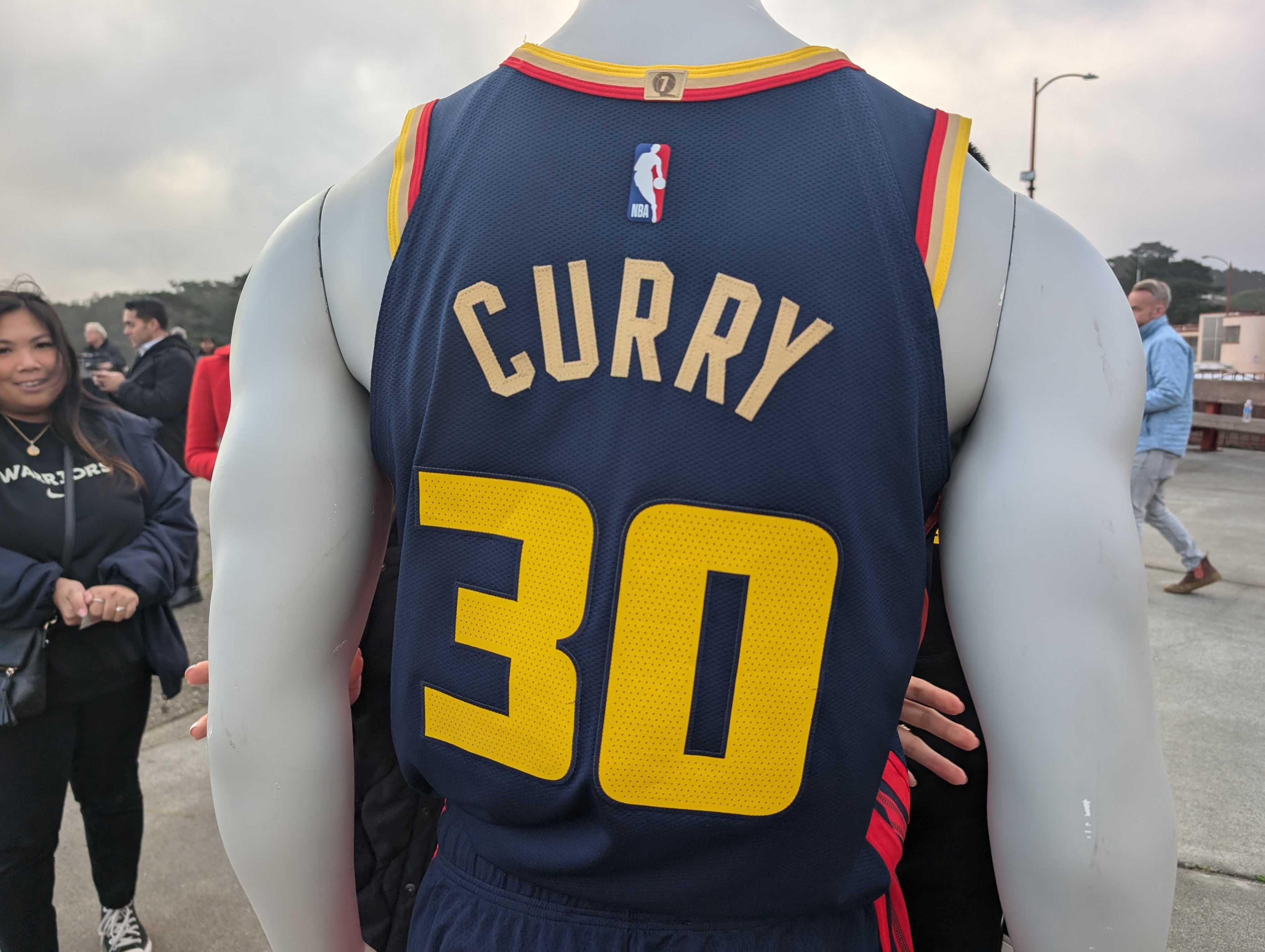 A mannequin wears a dark blue basketball jersey with &quot;Curry&quot; and the number 30 on the back. People in the background appear to be taking photos.