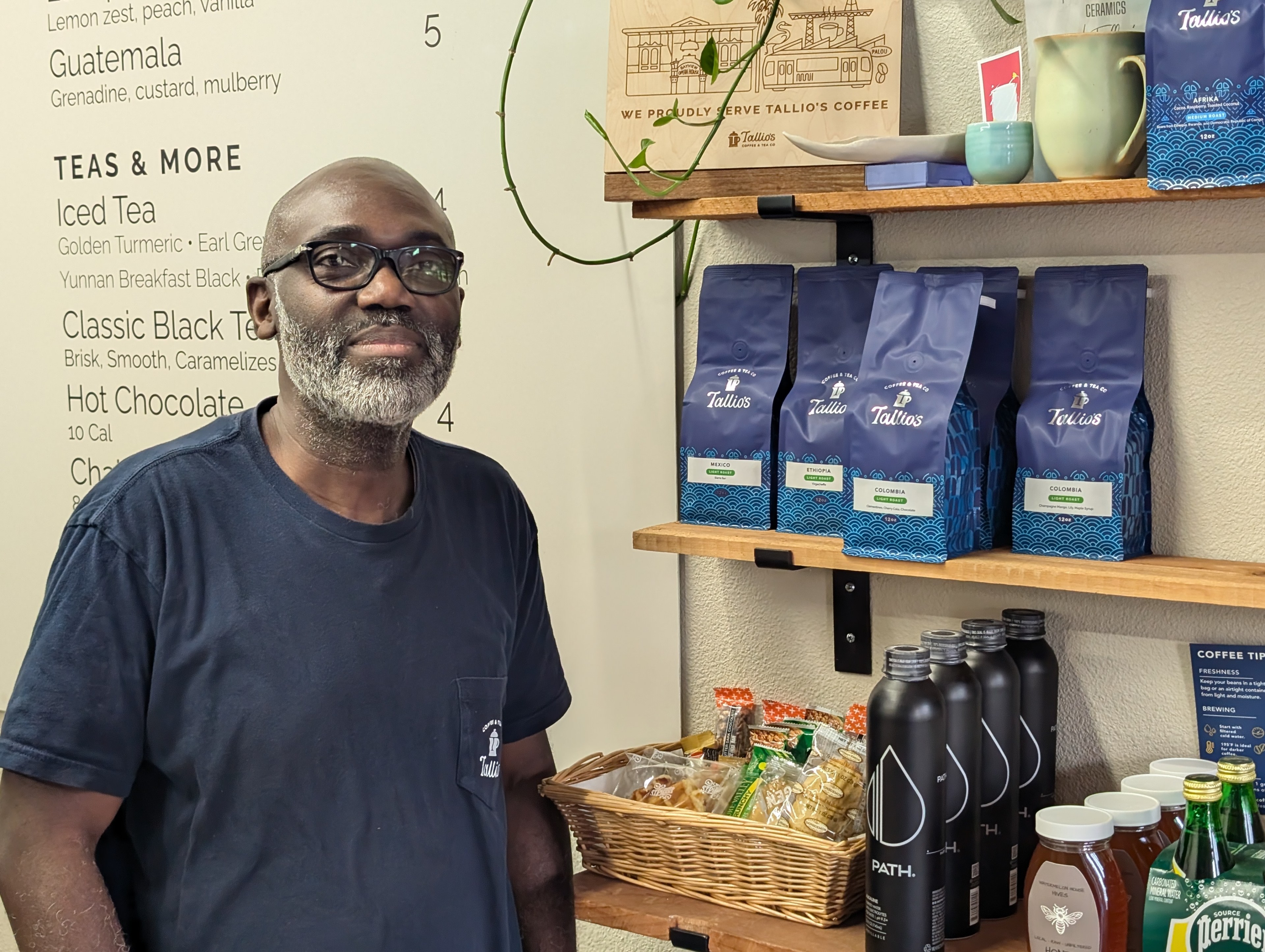 A person with glasses and a gray beard stands smiling by a shelf filled with bags of coffee labeled &quot;Tallio's,&quot; alongside snacks, drinks, and honey jars.