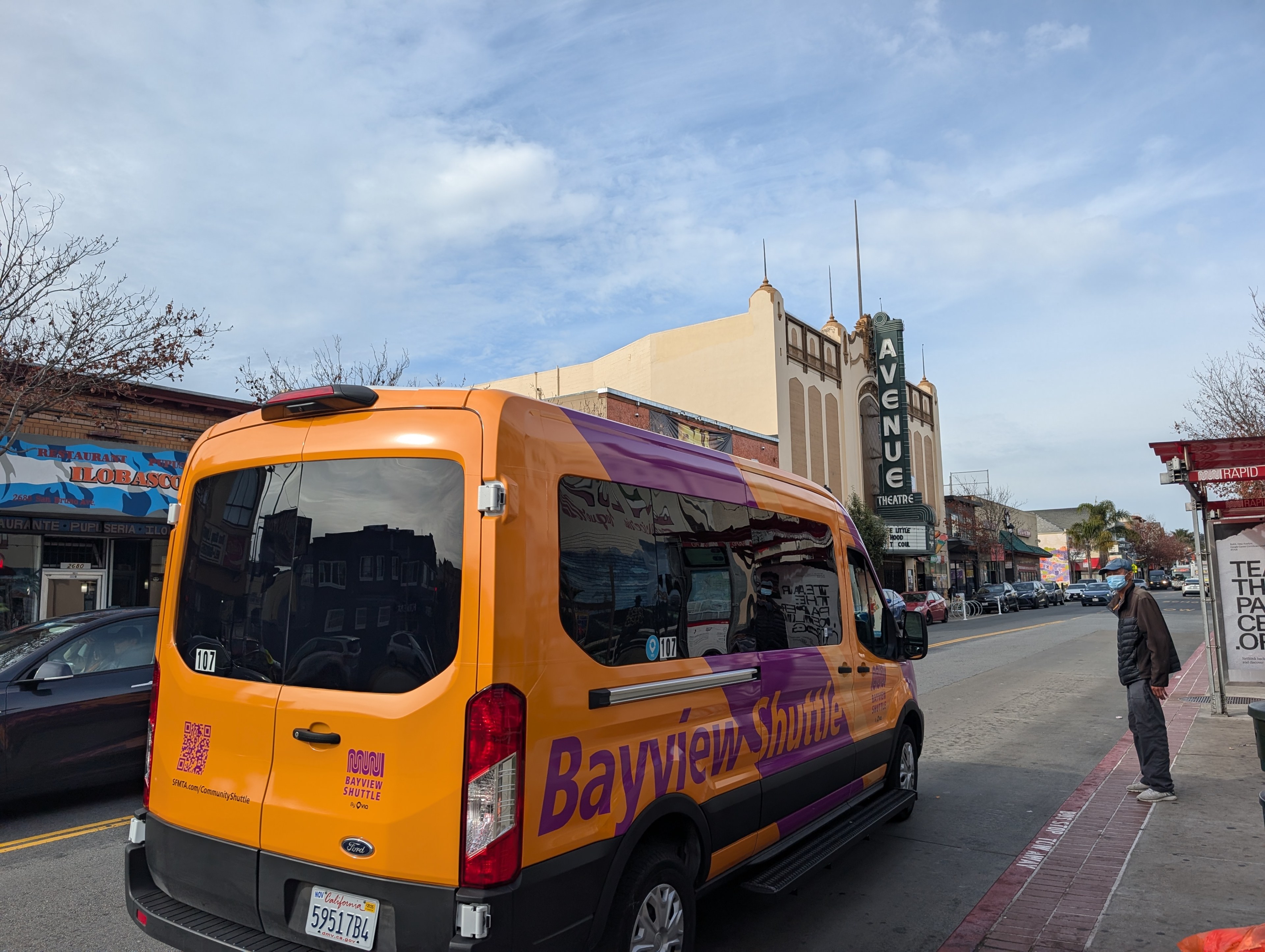 A bright orange shuttle van labeled &quot;Bayview Shuttle&quot; is parked on a street. Nearby, a person stands on the sidewalk, with a theater and shops visible in the background.