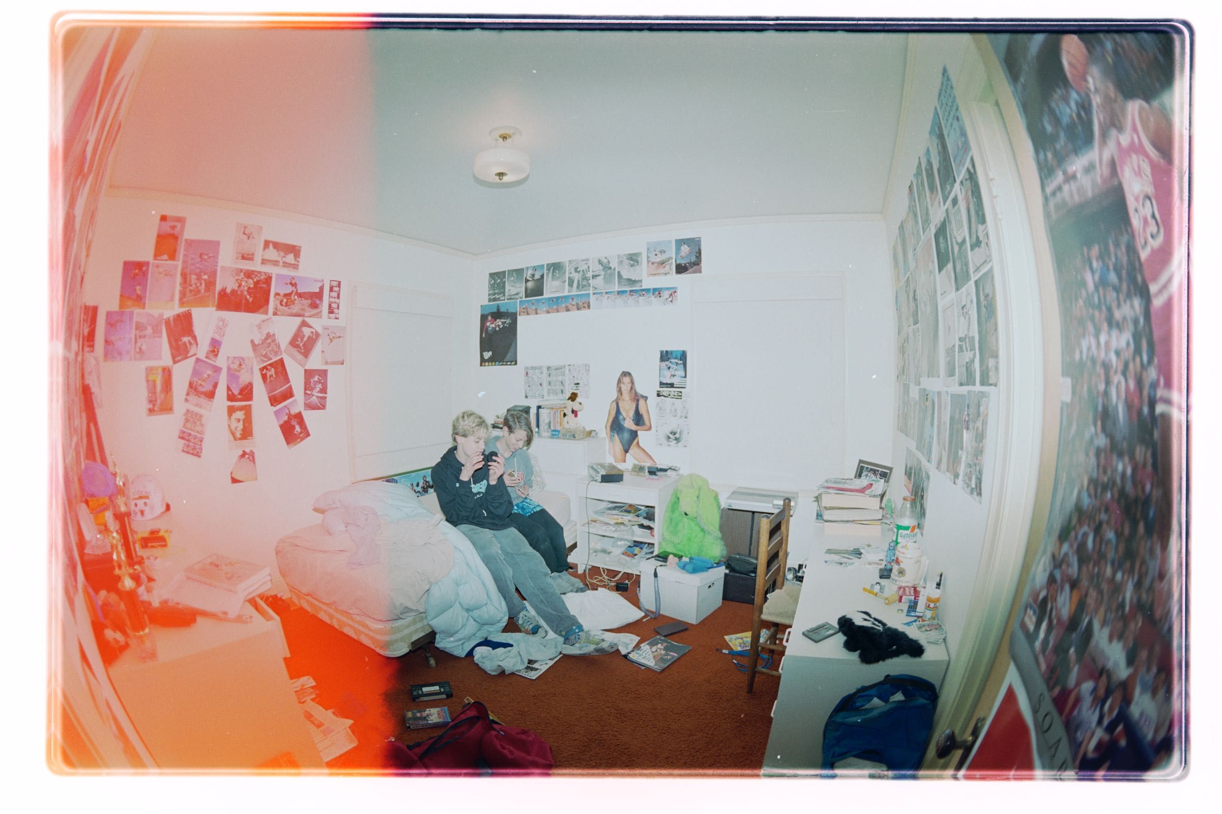 A cluttered room with posters on the walls and two people sitting on a bed. The room has a reddish hue, and there's a mix of personal items and furniture.