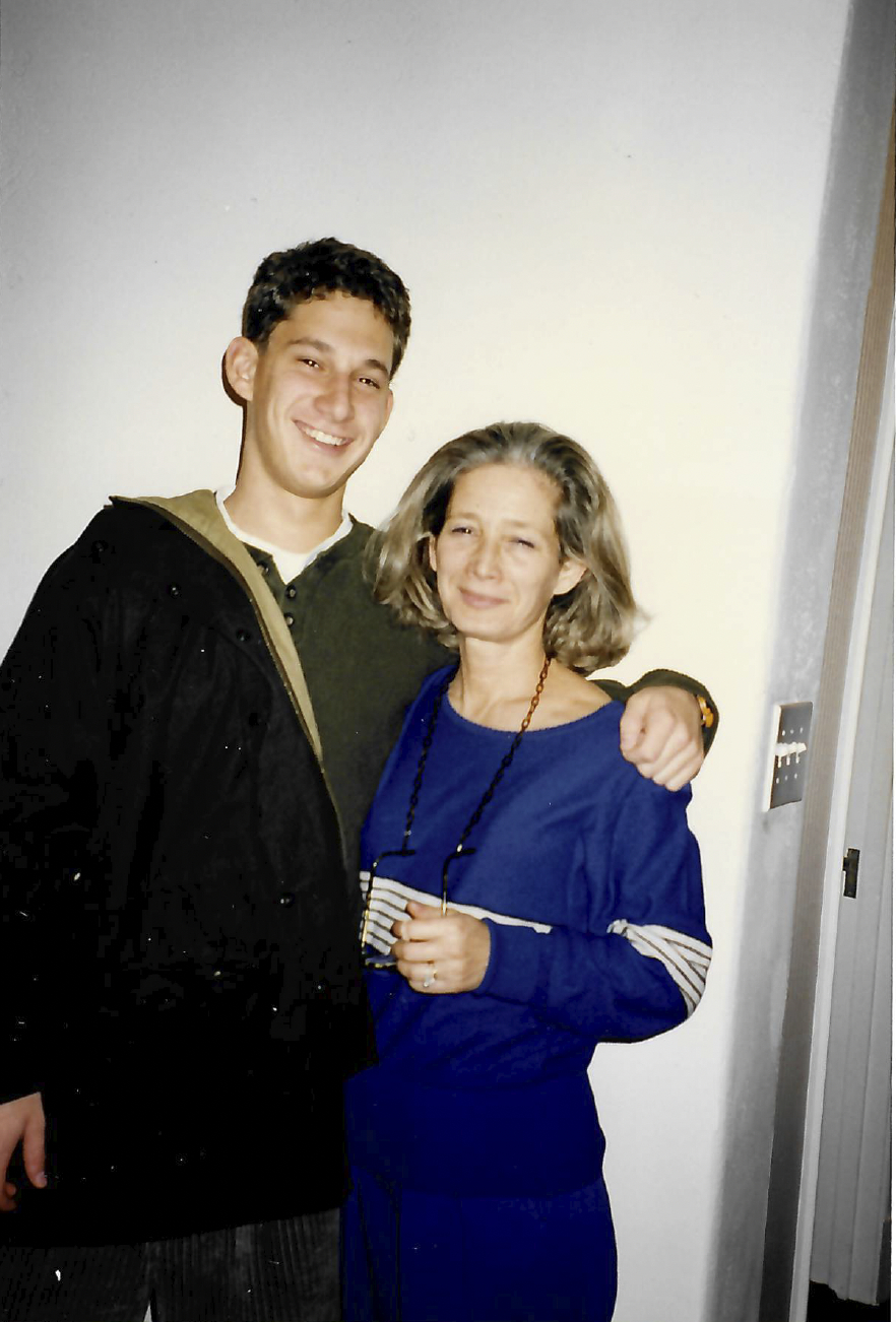 A young man and an older woman stand close together, smiling. The man wears a dark jacket, and the woman wears a blue outfit, holding glasses in her hand.