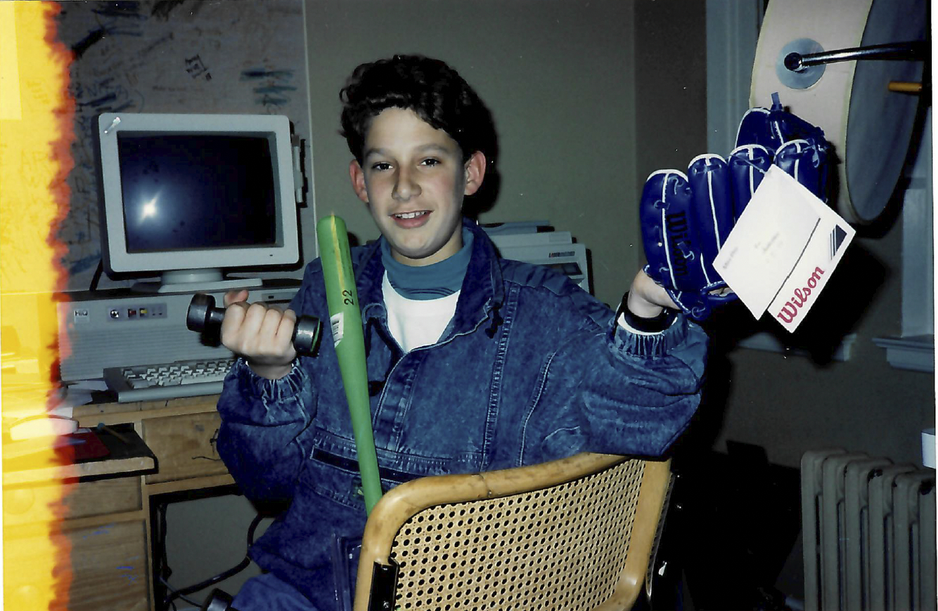 A young person in a denim jacket holds a blue baseball glove and a green bat, sitting in front of an old computer. The glove has a Wilson tag attached.