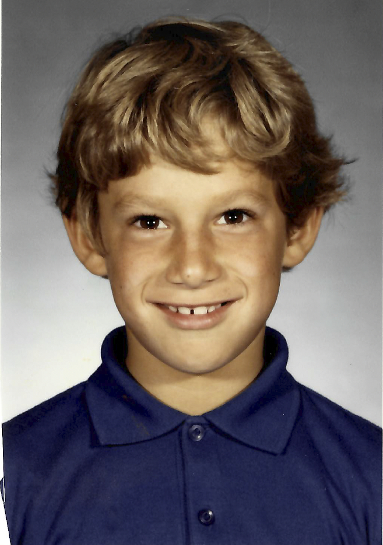 A young child with light brown hair smiles at the camera, wearing a blue collared shirt. The background is plain and softly lit.