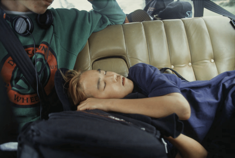 A person sleeps in the backseat of a car, wearing a blue shirt and resting on a black backpack. Another person sits beside them, wearing a green sweatshirt and headphones.