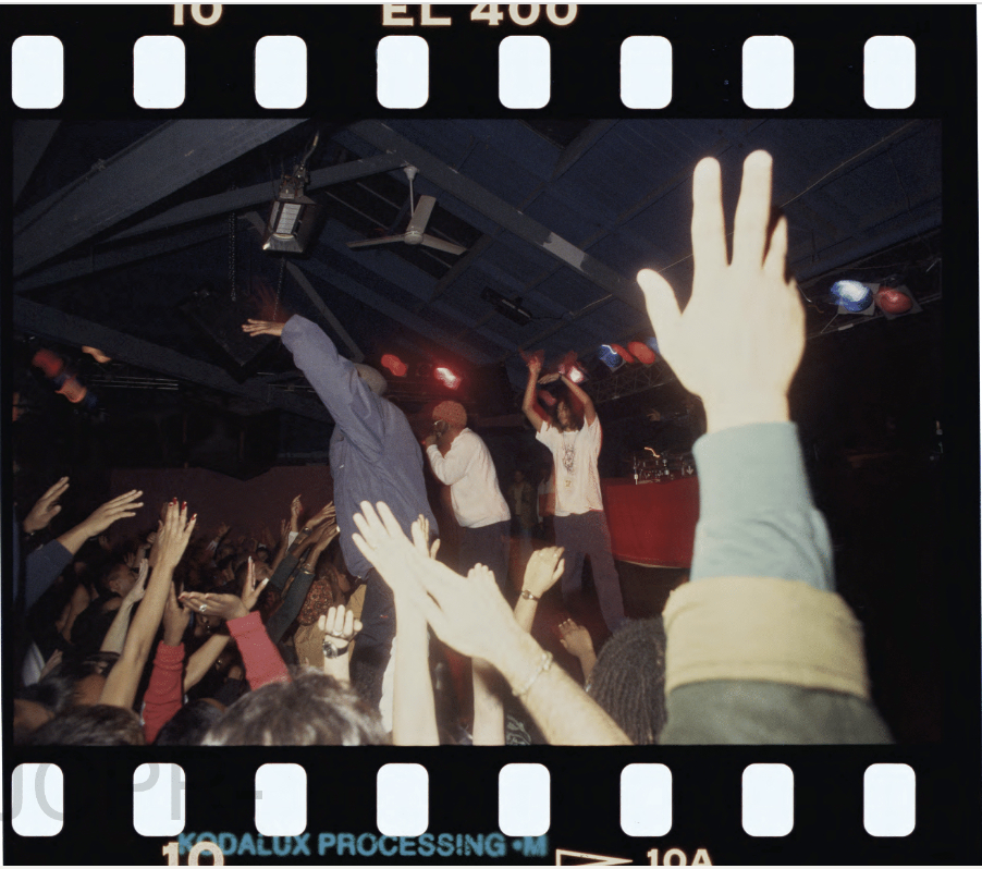 A lively concert scene shows two performers on stage energetically engaging with the crowd, whose hands are raised in excitement under dim, colorful lighting.