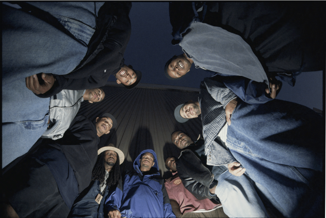 A group of nine people stand in a circle looking down at the camera, set against a tall, dark building with an overcast sky. They're casually dressed in jackets and hats.