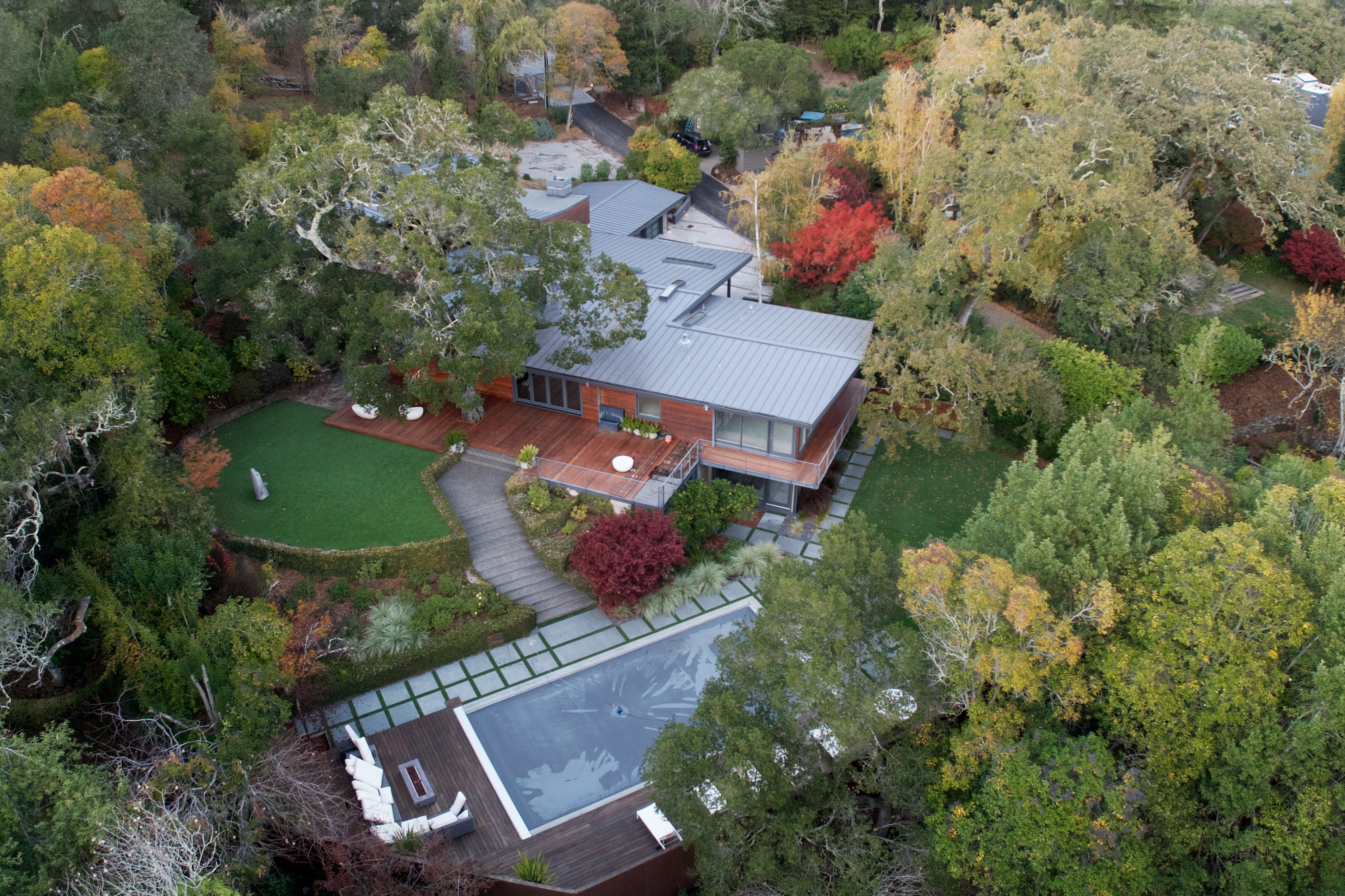 An aerial view of a modern house surrounded by dense greenery. It features a large pool, wooden decks, and landscaped gardens with a mix of colorful autumn trees.