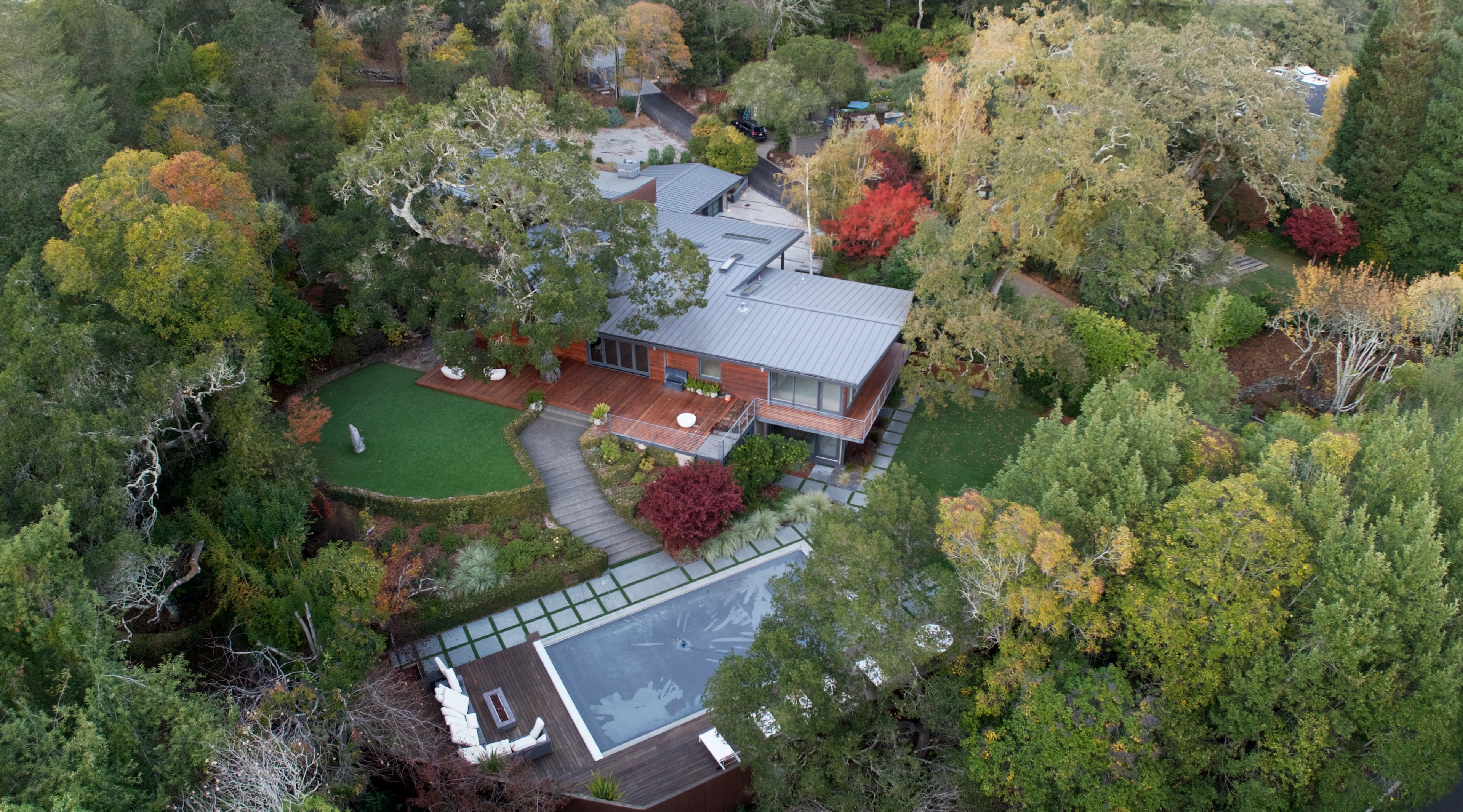 An aerial view of a modern house surrounded by dense greenery. It features a large pool, wooden decks, and landscaped gardens with a mix of colorful autumn trees.