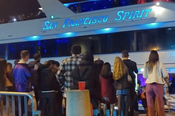 A crowd stands near a docked ship named "San Francisco Spirit" at night, with blue lights illuminating the vessel. A red cup is on a nearby concrete bollard.