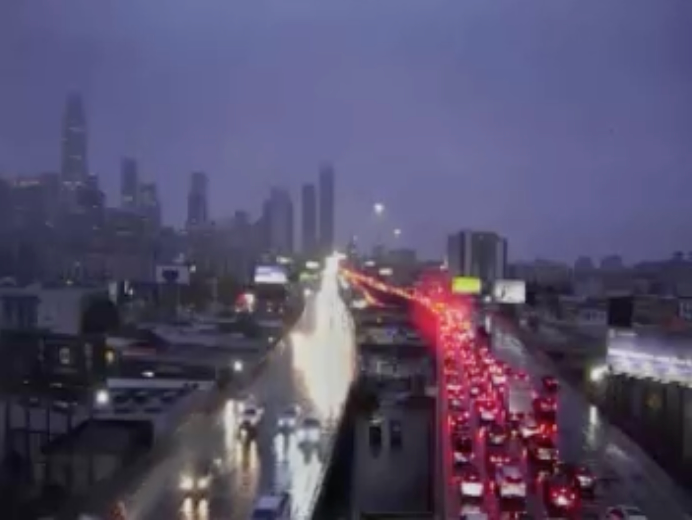 A rainy cityscape with heavy traffic, featuring wet reflecting roads. Red taillights glow on one side, skyscrapers fade into a cloudy, gray-blue sky.