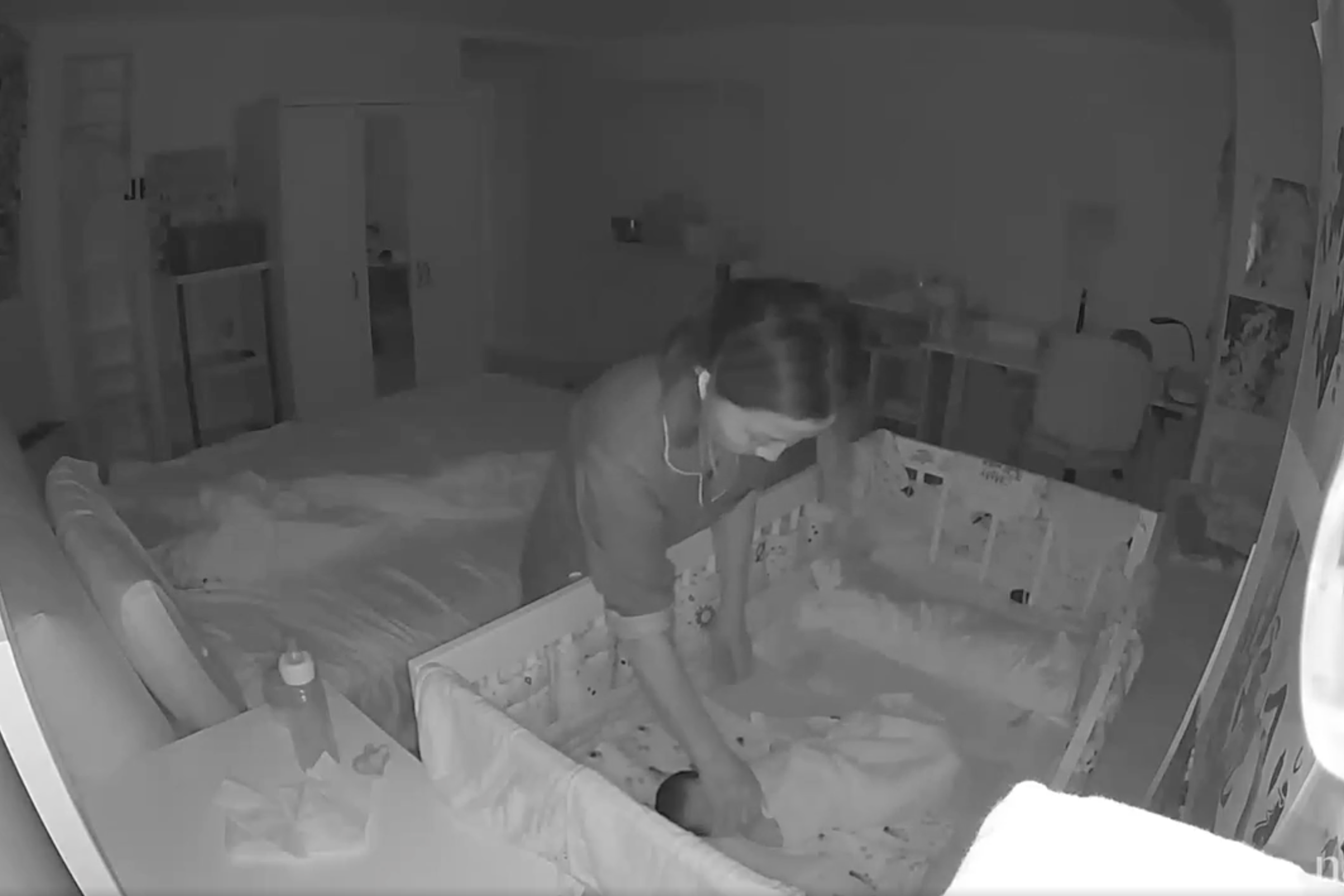 A woman is bending over a crib, attending to a baby. The scene is dimly lit, likely by night vision, showing a bedroom with a bed and a few pieces of furniture.