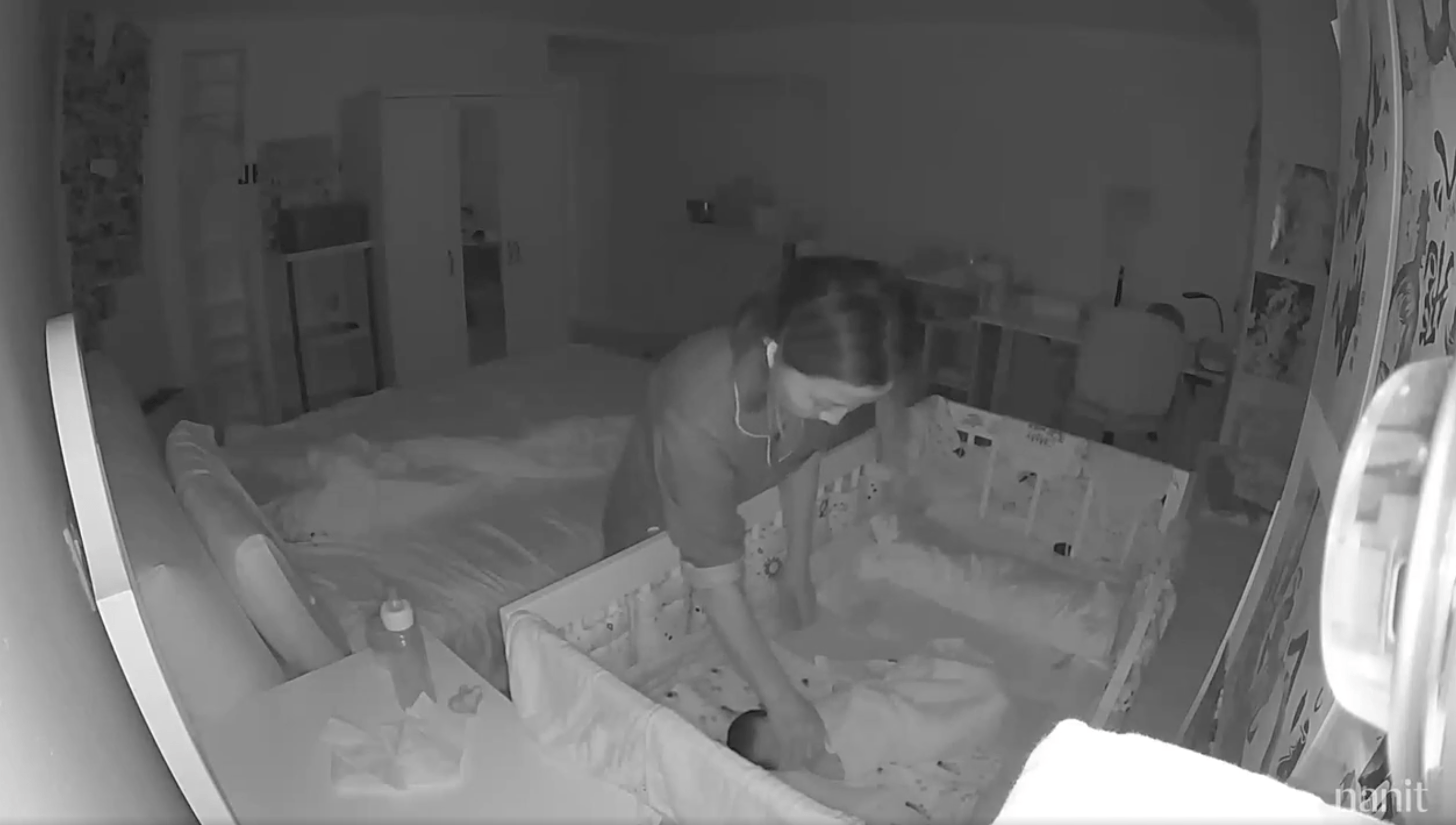 A woman is bending over a crib, attending to a baby. The scene is dimly lit, likely by night vision, showing a bedroom with a bed and a few pieces of furniture.