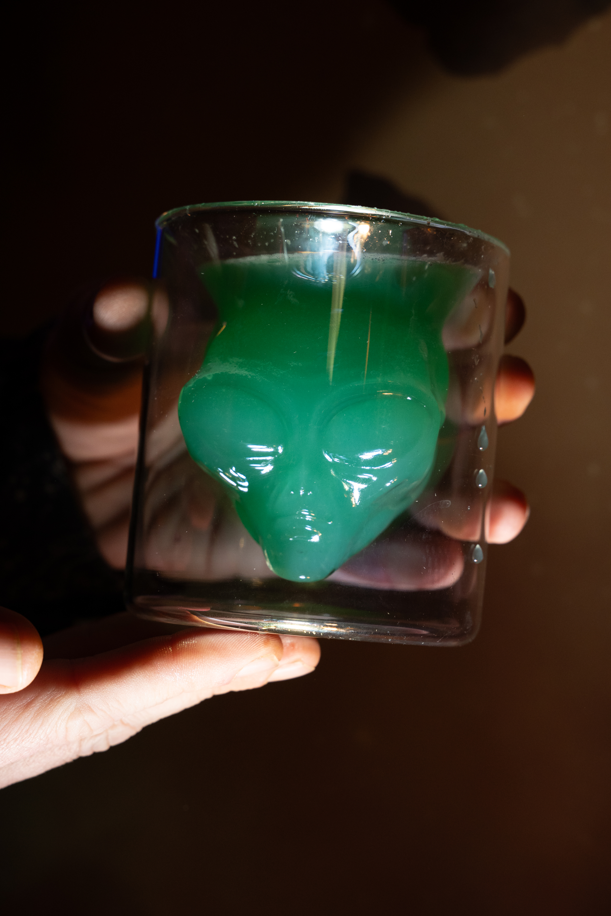 A hand holds a glass shaped like an alien face containing a green cocktail. The glass is slightly reflective under light.