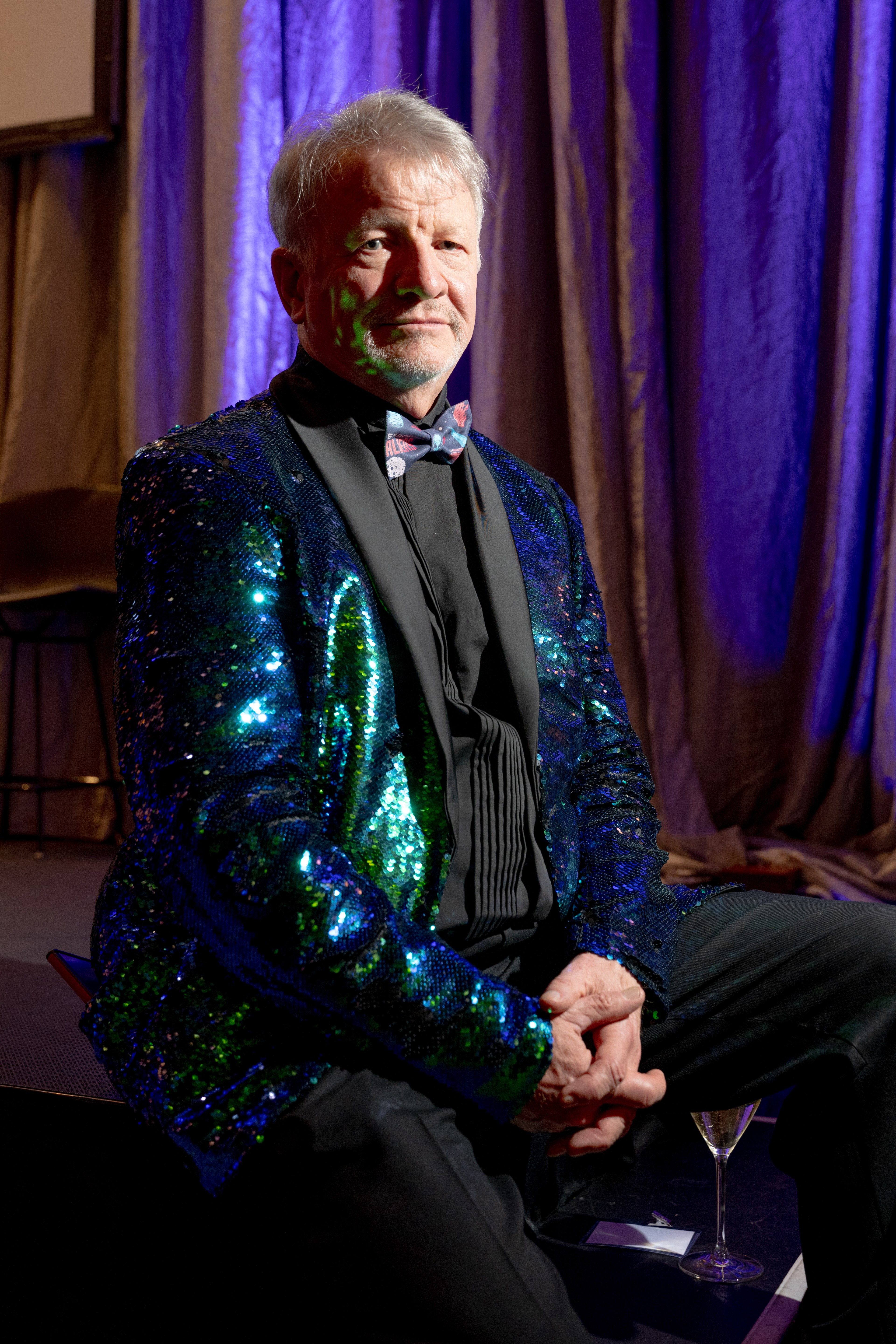 A man is seated with a confident expression, wearing a shimmering blue and green sequin jacket and a bow tie, against a backdrop of purple curtains.