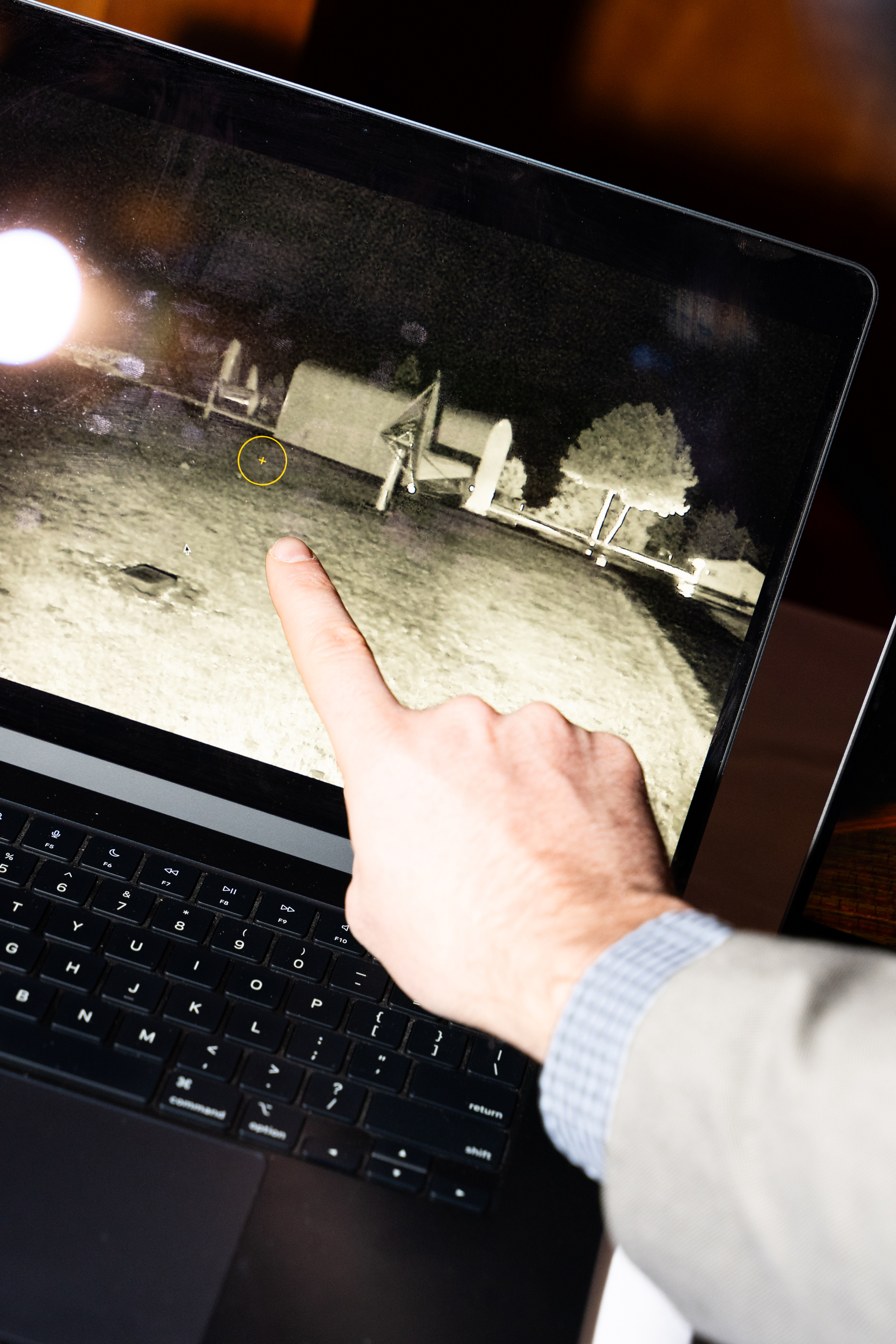 A person points at a black and white image on a laptop screen showing outdoor equipment and trees. A circled area draws focus on the screen.