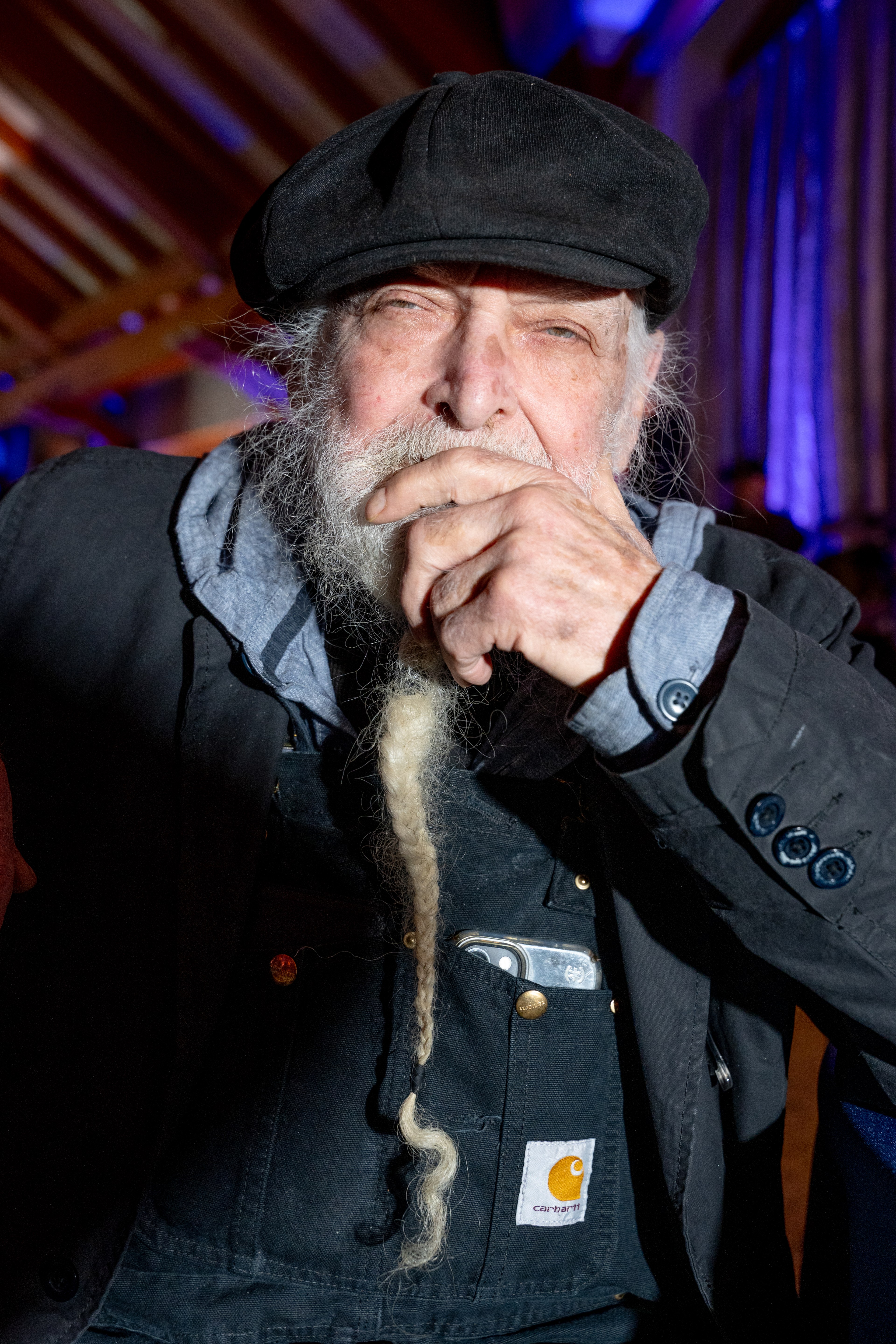 An elderly man with a long, braided beard wears a black cap and jacket, resting his chin on his hand. The background is softly lit in blue.