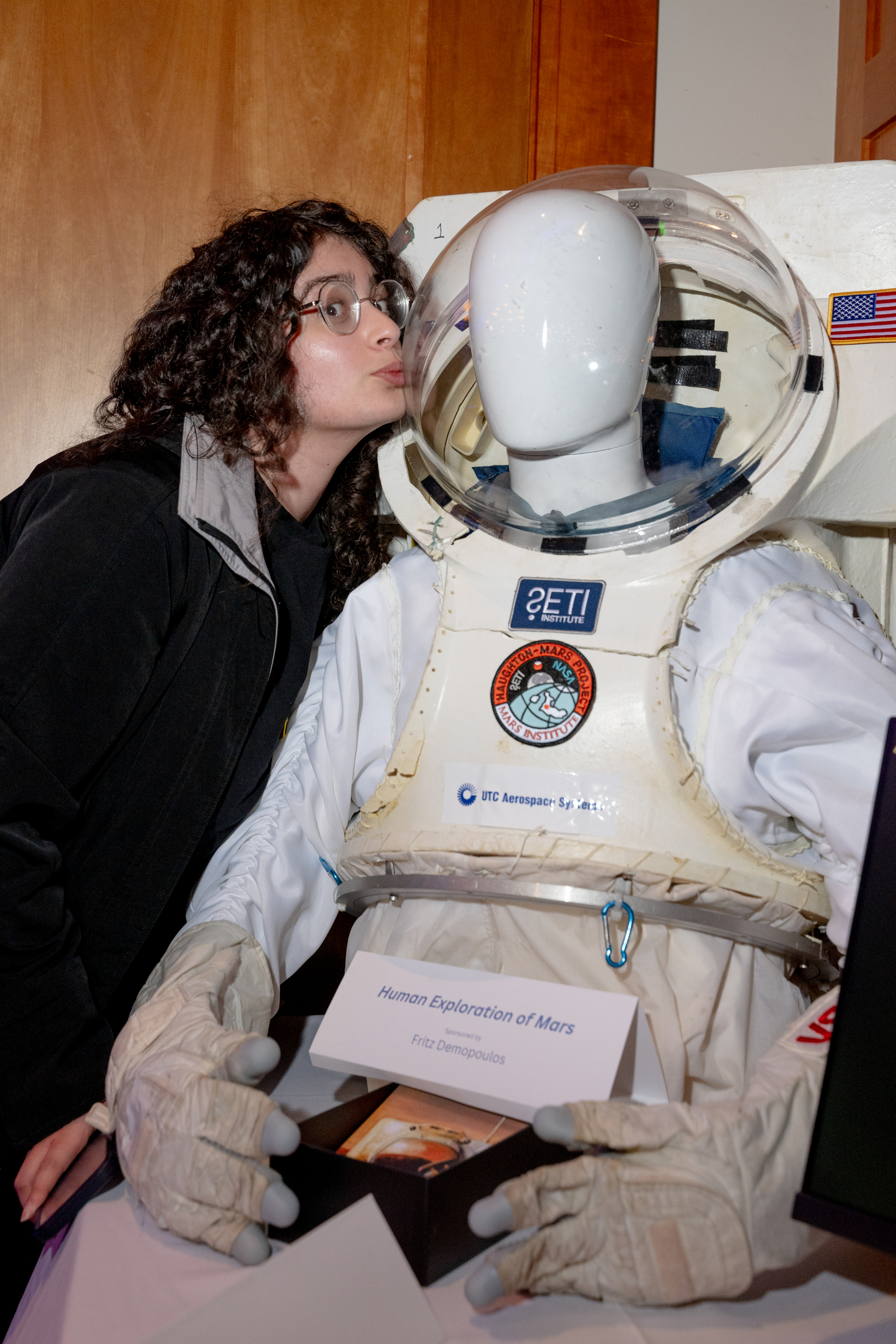 A person playfully kisses the helmet of a mannequin dressed in a space suit with various logos and a sign titled &quot;Human Exploration of Mars&quot; nearby.