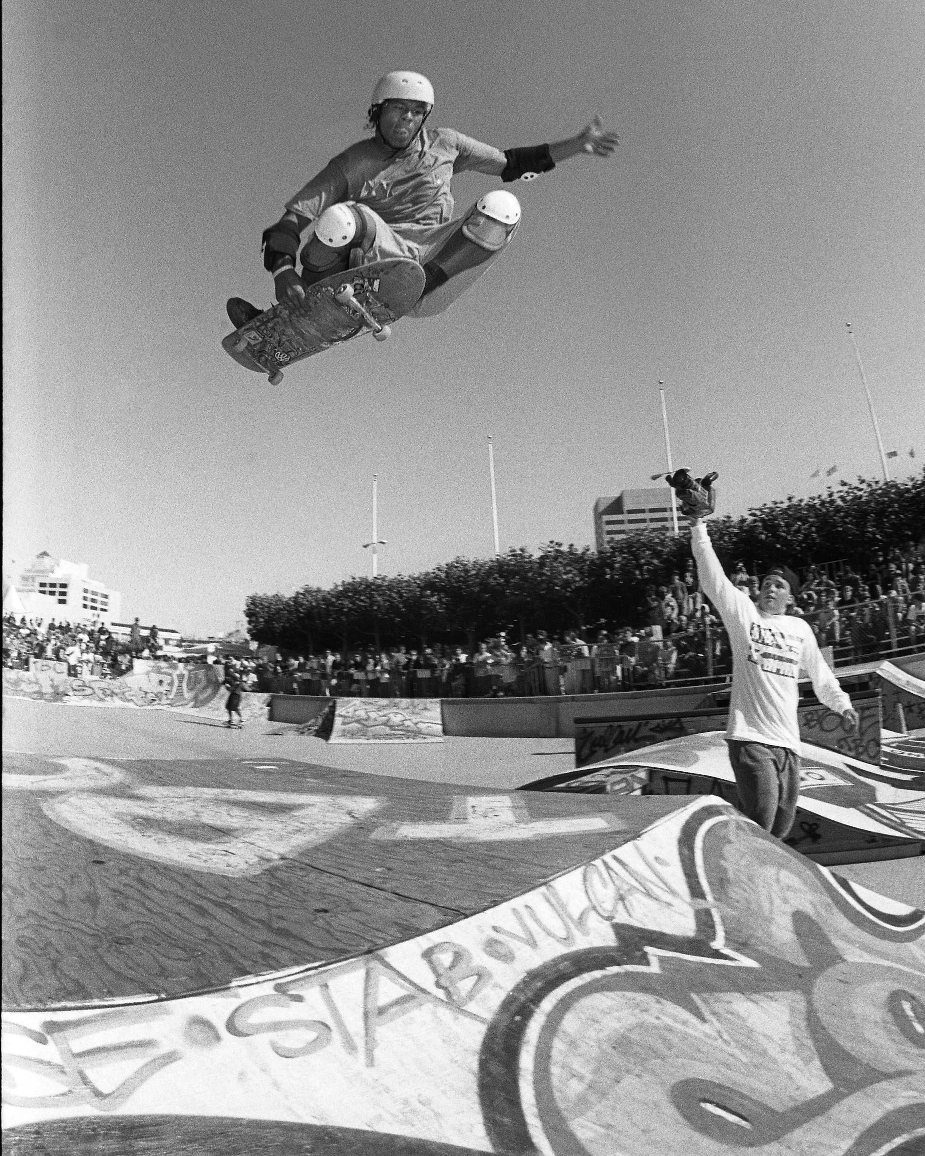 A skateboarder performs an aerial trick, wearing a helmet and pads. A cheering spectator watches, and graffiti decorates the skate park in the background.