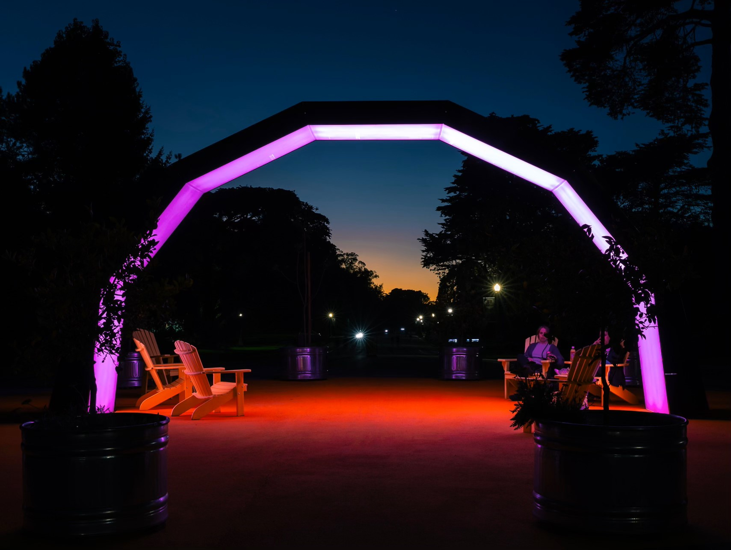 A glowing pink archway at dusk lights up a few wooden chairs and two people beneath it, creating a cozy atmosphere, framed by dark trees.