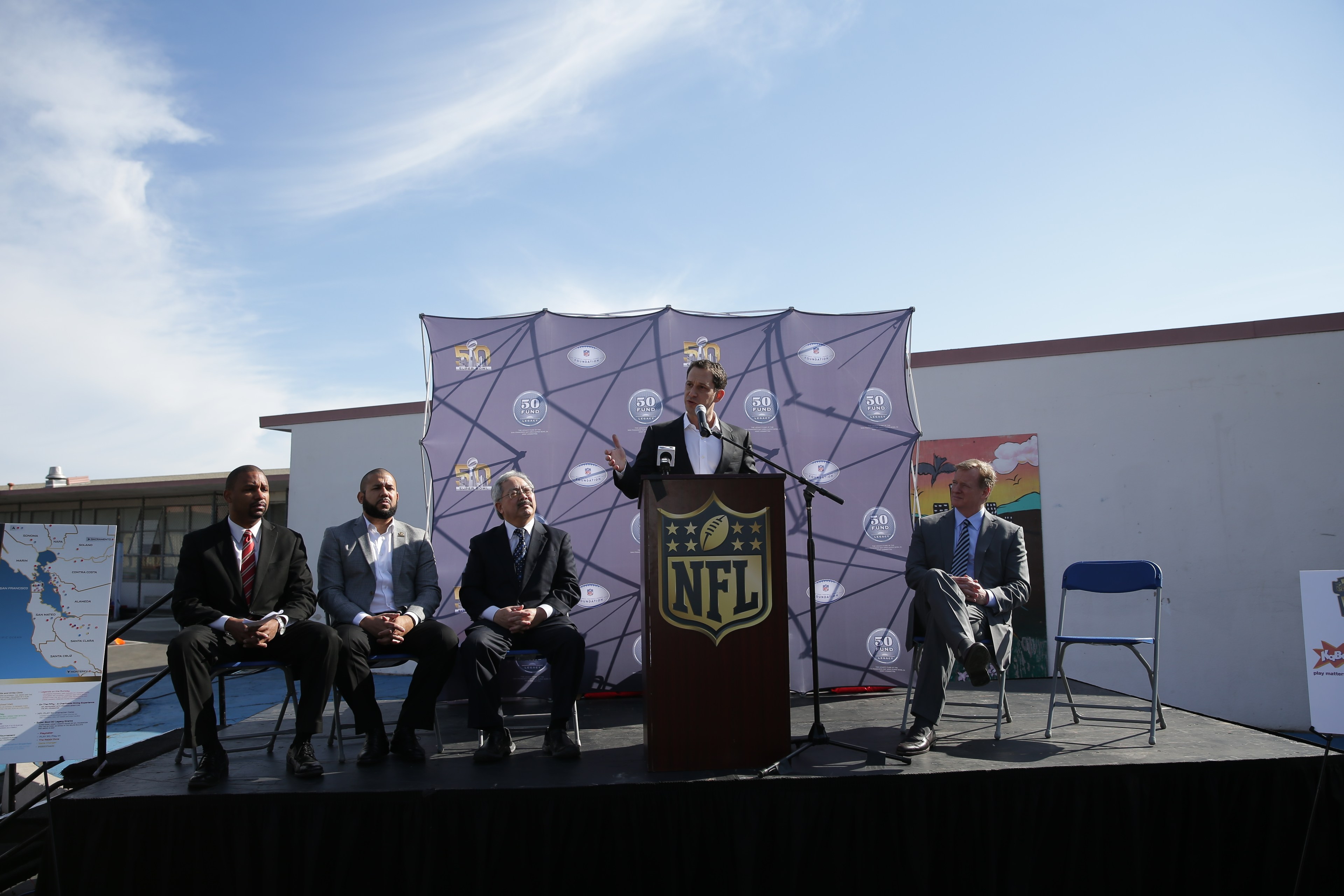 A man speaks at a podium with an NFL logo, flanked by four seated men in suits. They are outdoors with a blue sky backdrop and an NFL-themed banner.