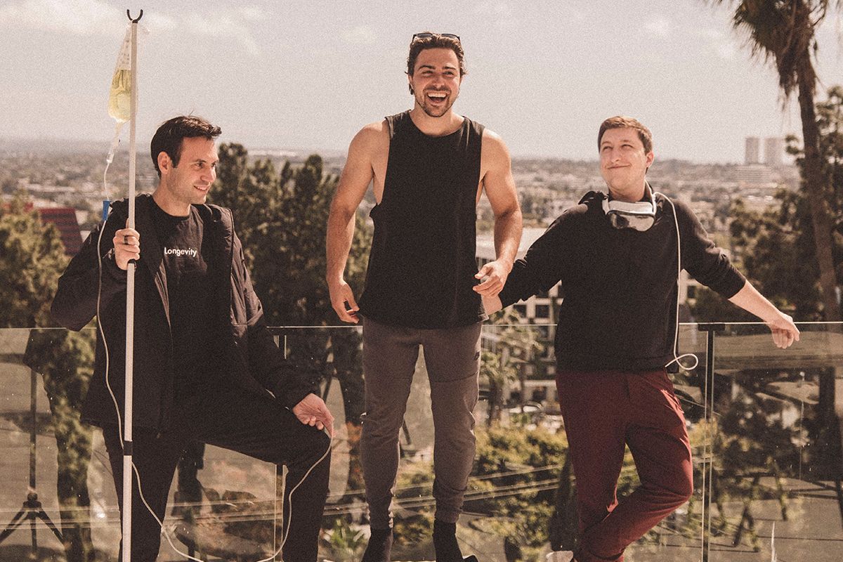 Three men are on a sunny balcony. One is sitting while holding a pole, another is standing on a mini trampoline, and the third is standing with headphones.