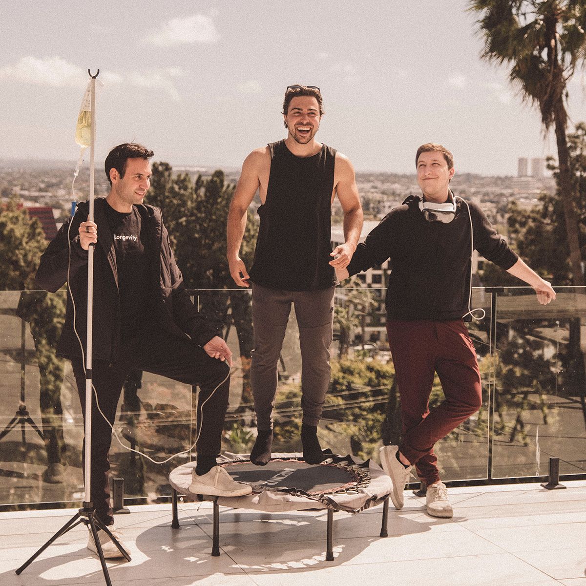 Three men are on a sunny balcony. One is sitting while holding a pole, another is standing on a mini trampoline, and the third is standing with headphones.