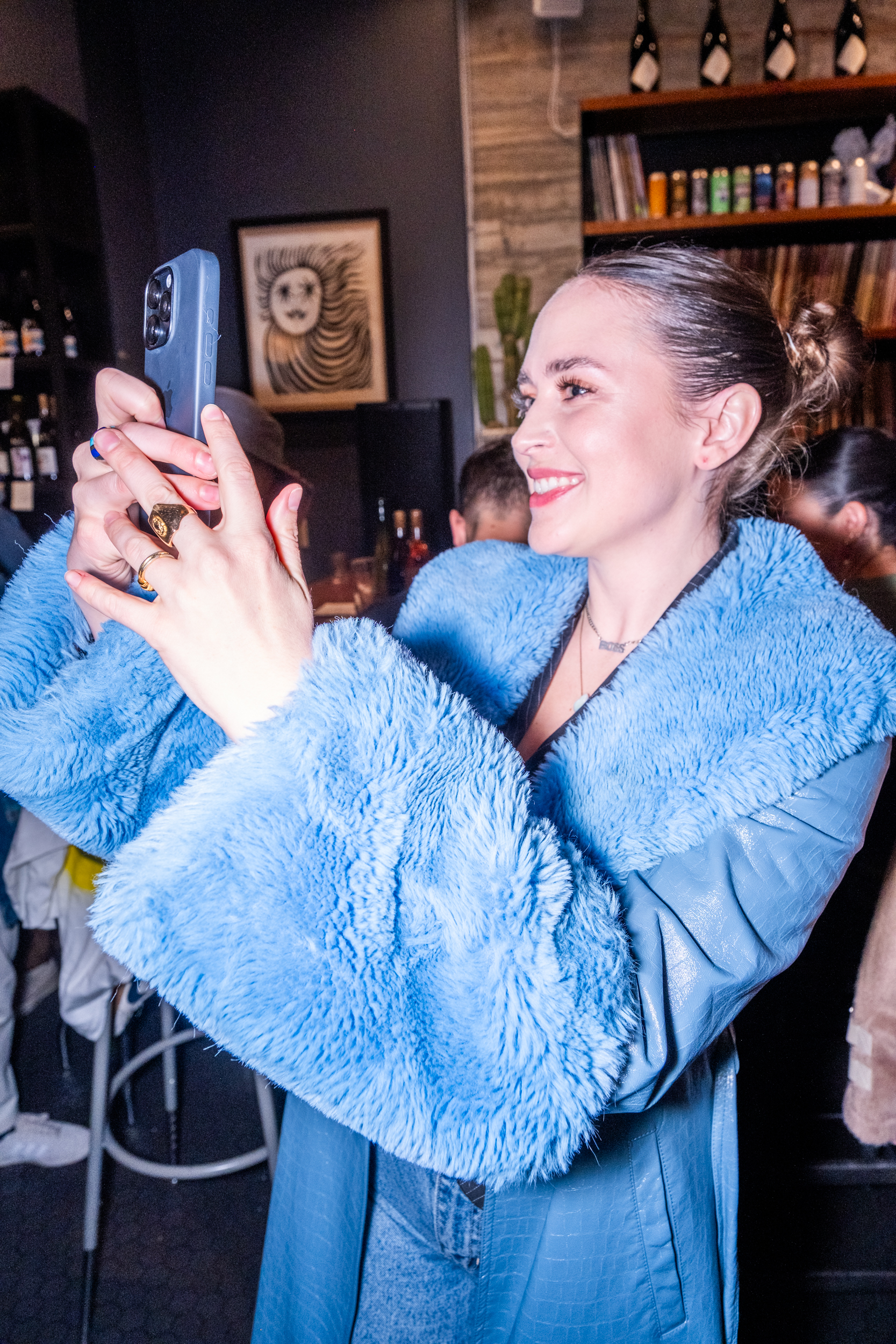 A woman in a bright blue faux fur coat smiles as she takes a photo with her phone. The background shows a bar setting with bottles on shelves.