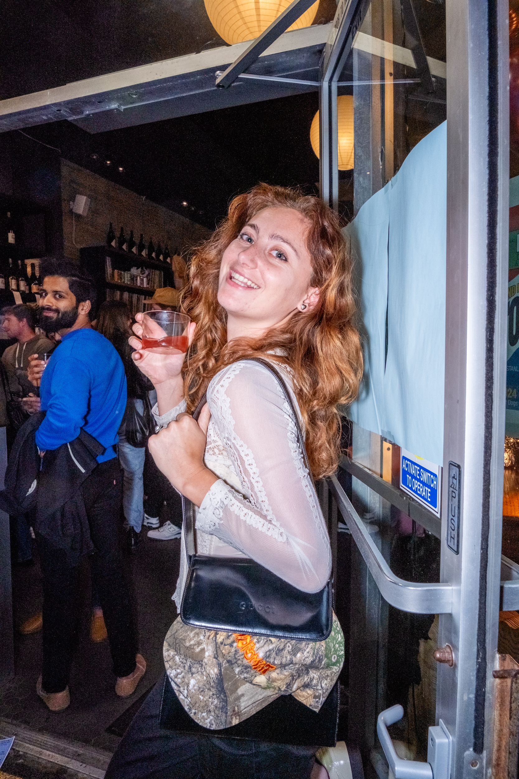 A woman with reddish hair, smiling, holding a drink, stands in a doorway at a lively bar. People in the background are socializing under warm, spherical lights.